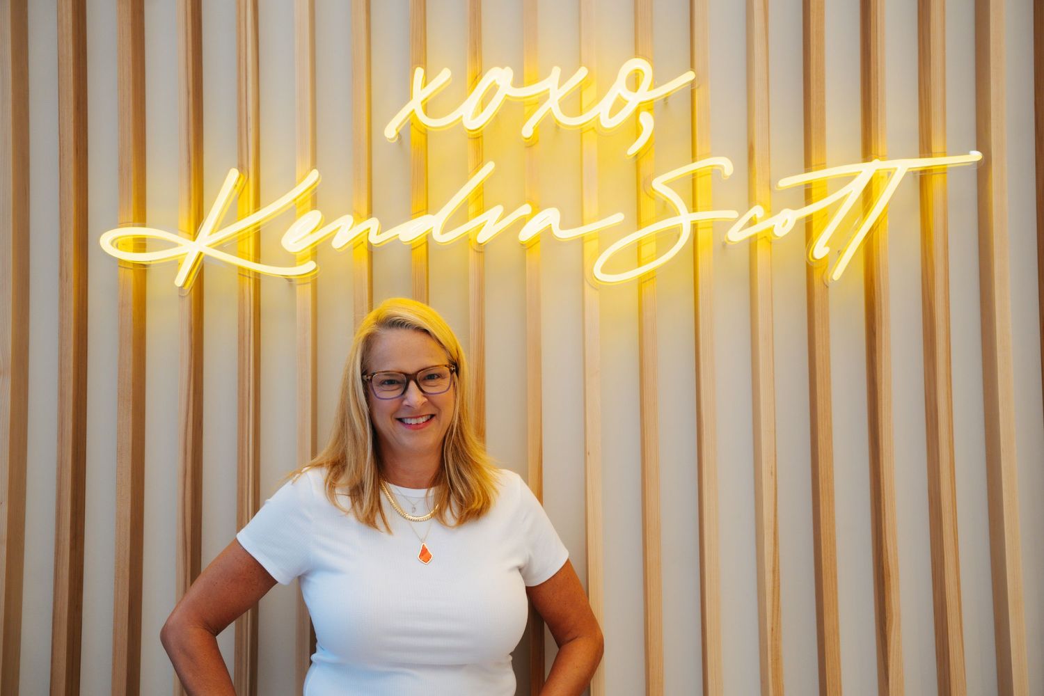 Blonde woman in white top with orange necklace before XOXO Kendra Scott sign.