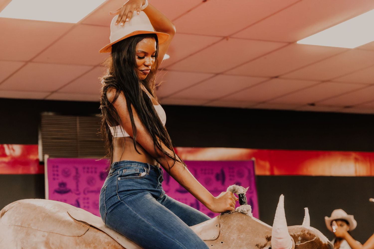 Woman in orange cowboy hat rides a mechanical bull indoors.