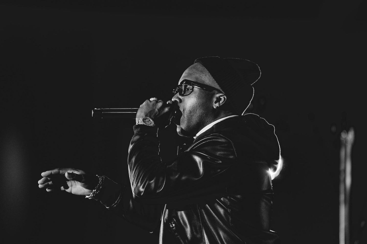 Black and white photo of a male performer singing into a microphone on stage wearing glasses and a leather jacket.