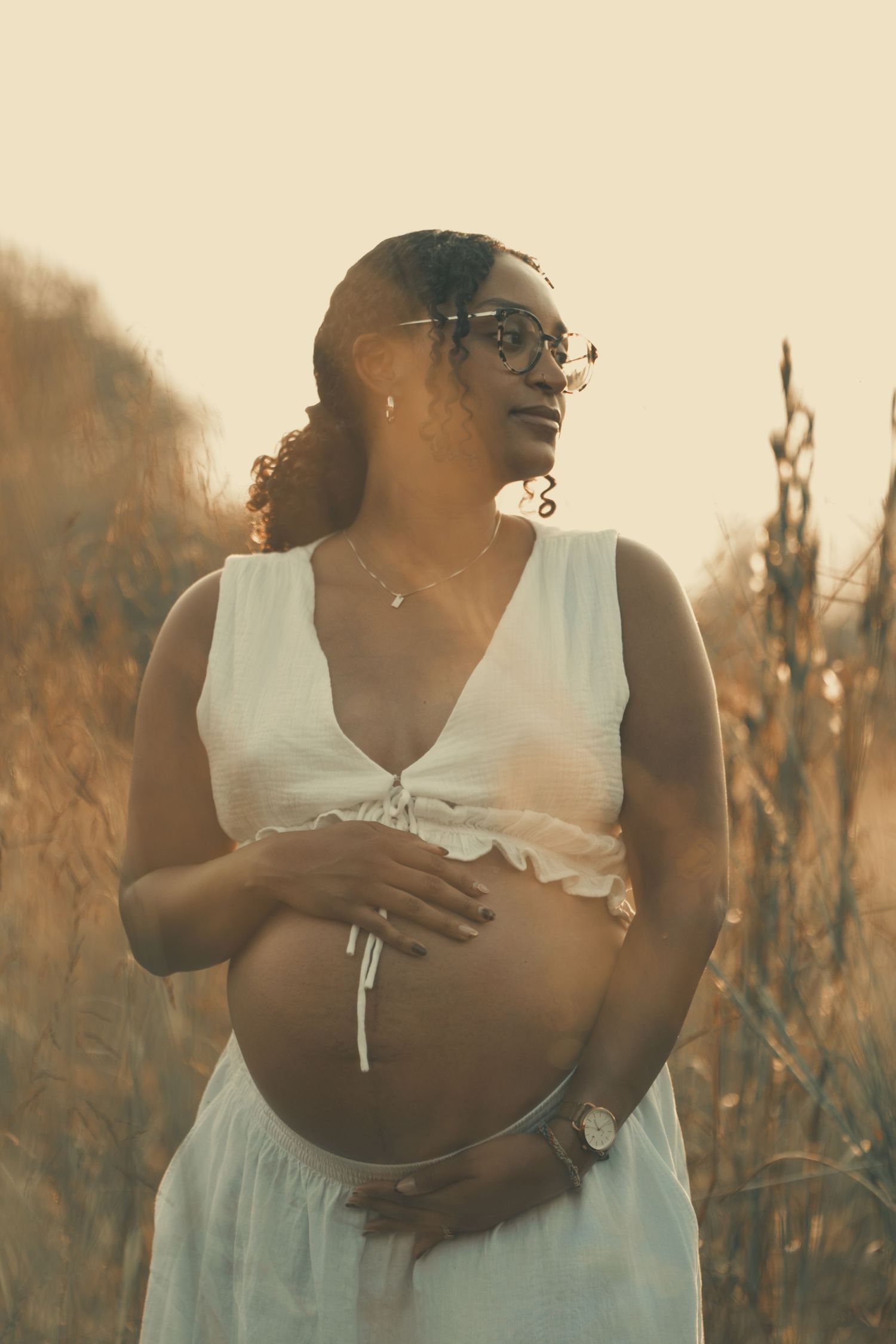 Smiling pregnant woman in white dress holds belly in golden field at sunset.