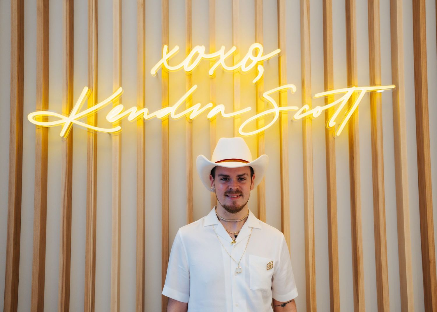 Man in white outfit and cowboy hat posing before XOXO Kendra Scott neon sign.