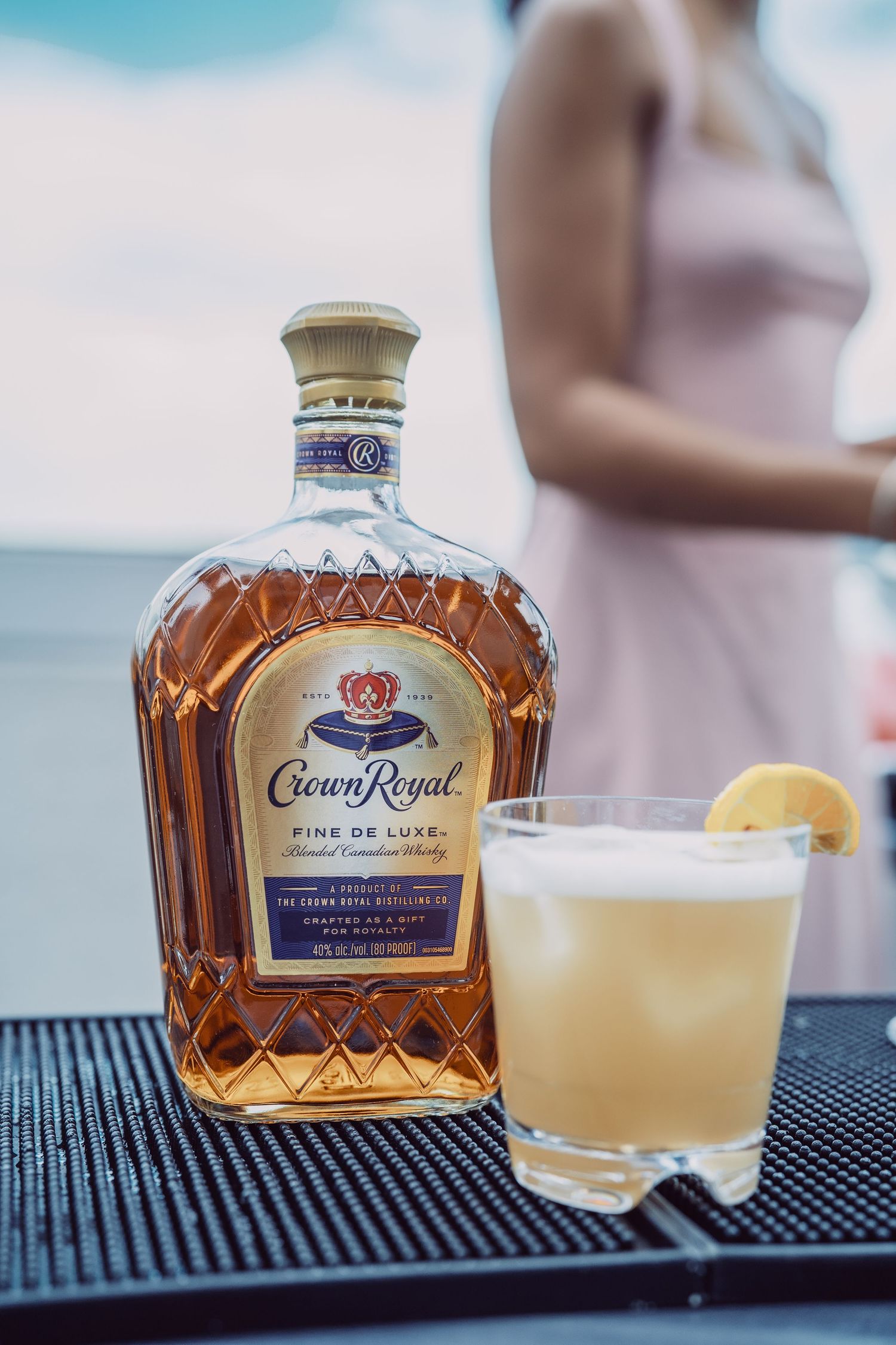 Crown Royal Fine De Luxe whisky bottle beside a lemon cocktail on a bar.