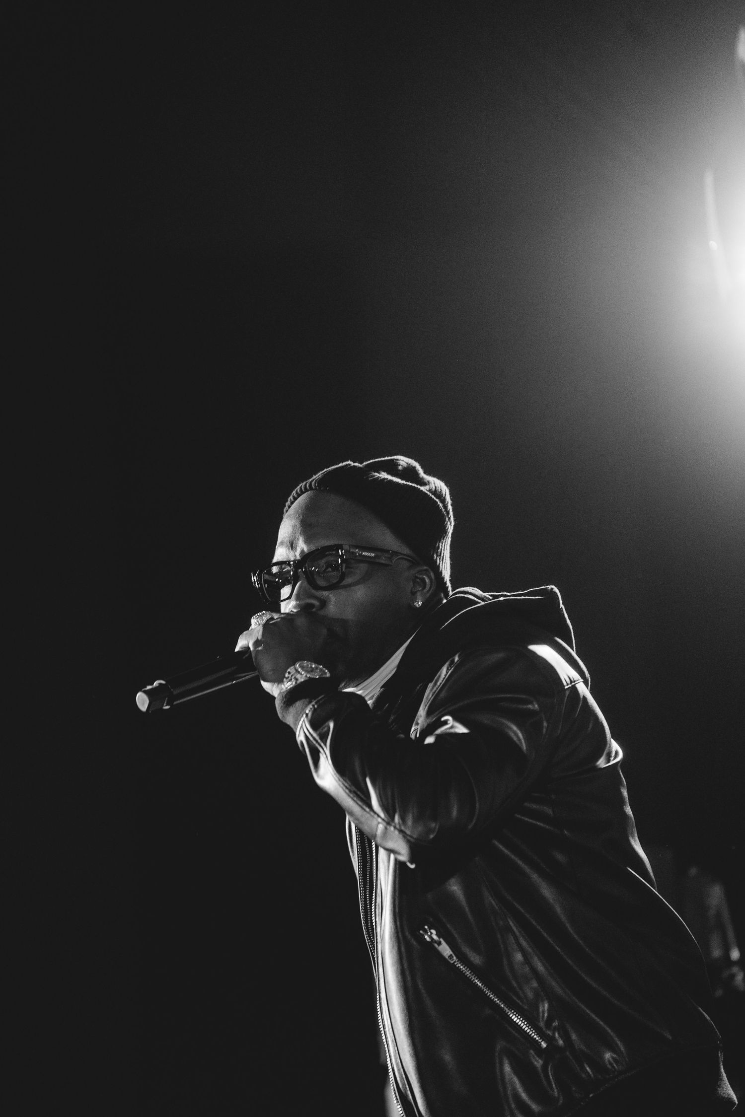 Black and white dramatic concert photo of male rapper performing with microphone under a single spotlight.