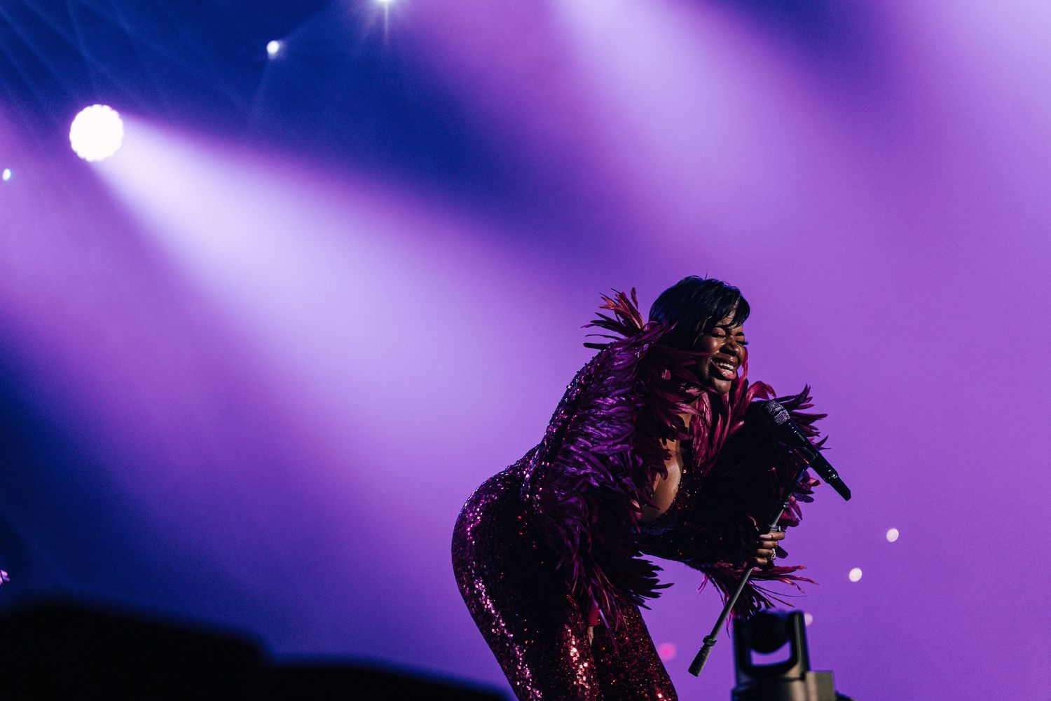 Female performer in sparkling feathered outfit singing passionately on stage under vibrant purple concert lighting.
