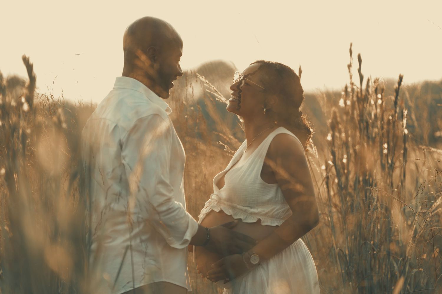 Couple shares joyful moment in sunlit field during maternity photoshoot.