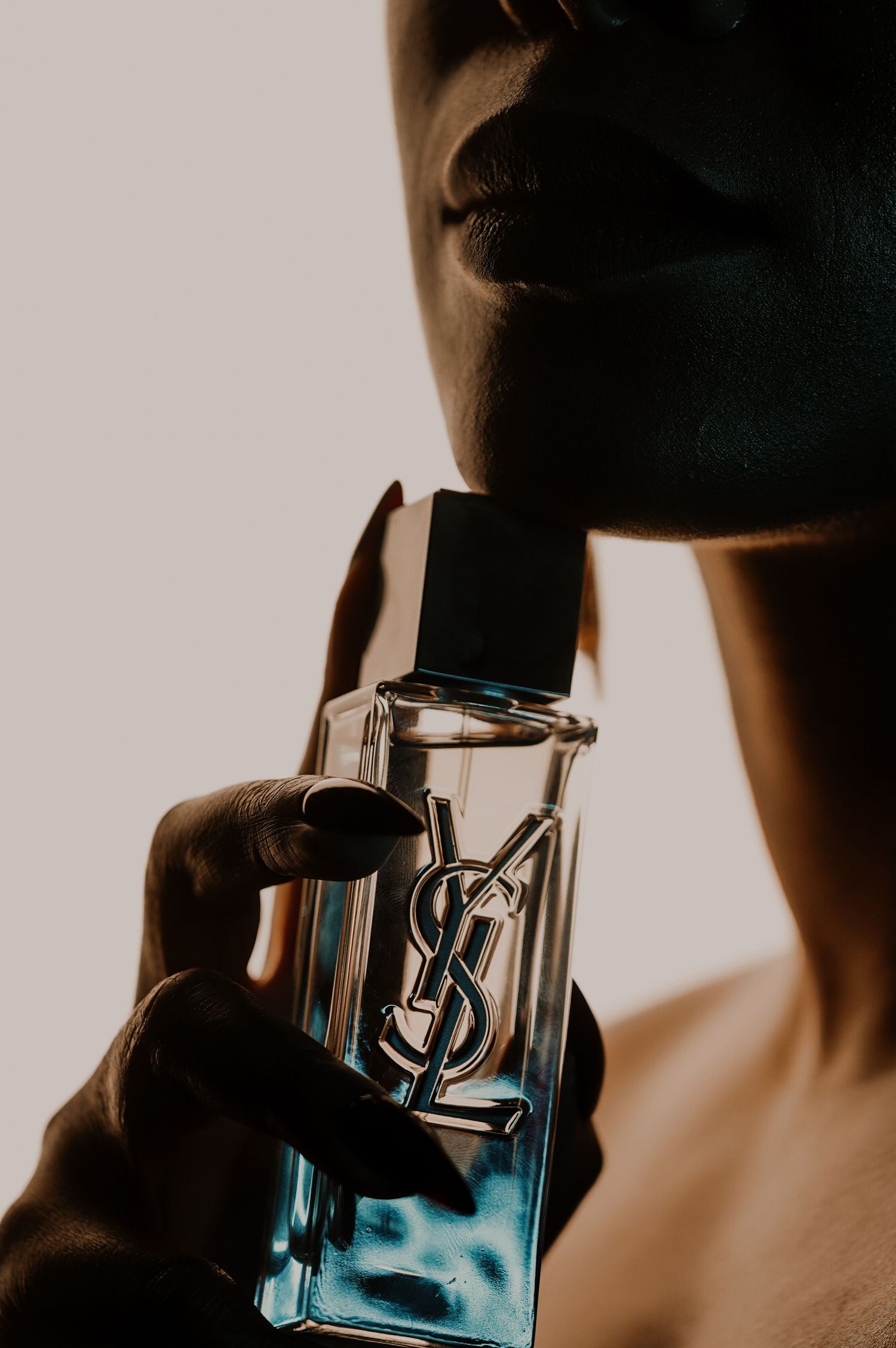 Silhouetted woman with dark manicured nails holding a blue YSL perfume bottle against her chin in warm, moody backlighting.