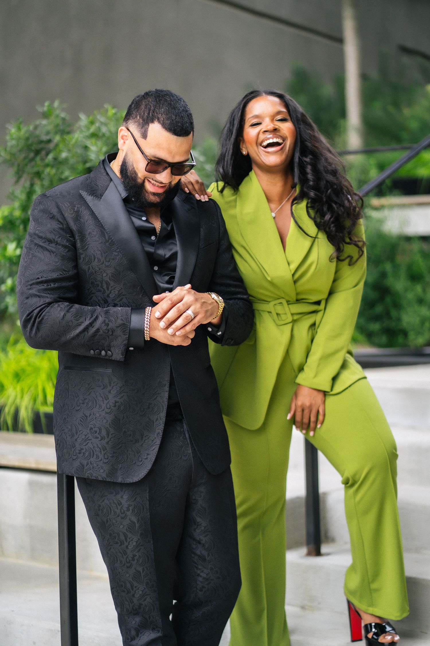 Stylish couple laughing together, man in black suit and woman in green pantsuit outdoors.