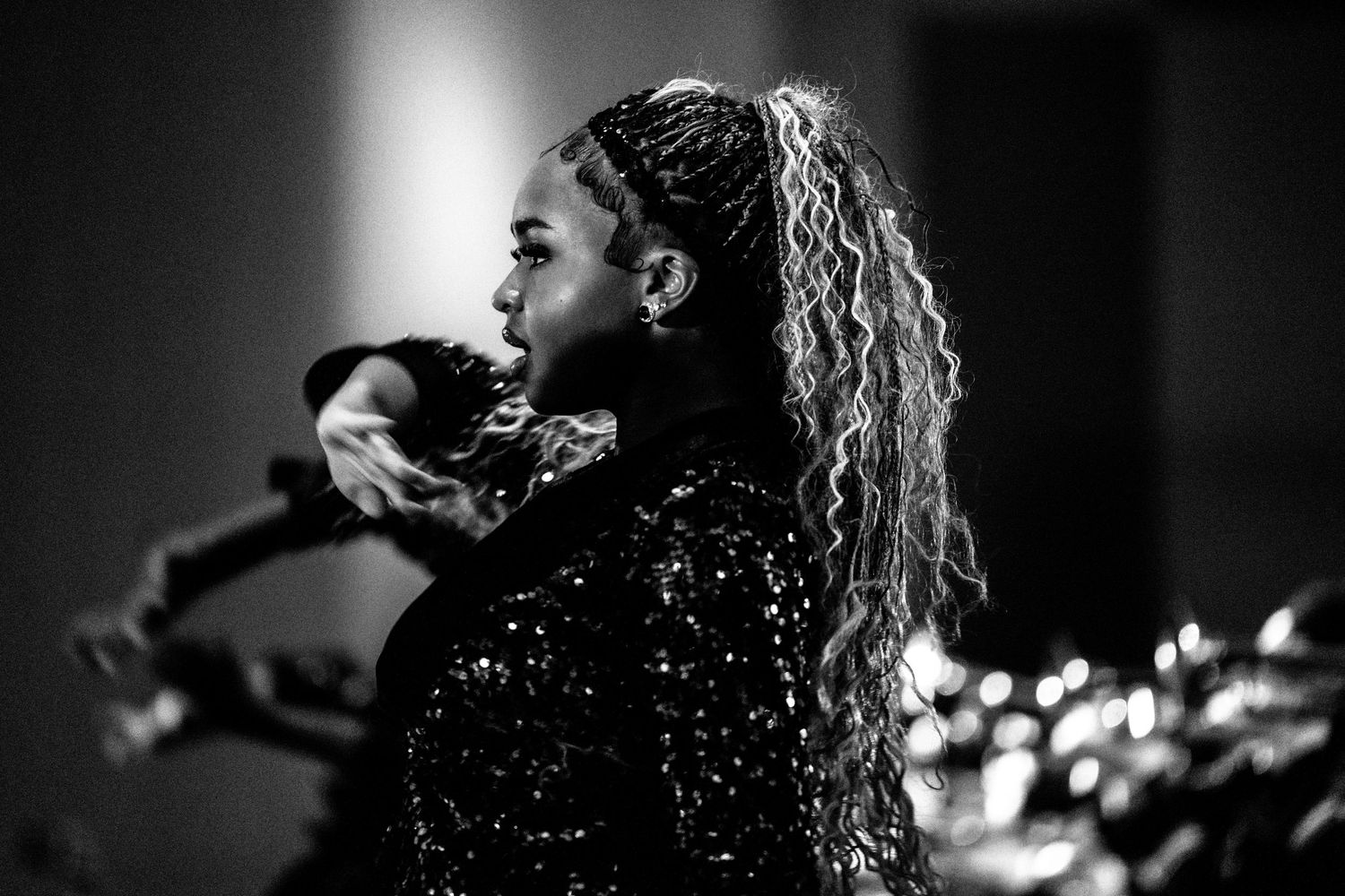 Black and white profile shot of female performer with long curly ponytail in sequined outfit on stage.