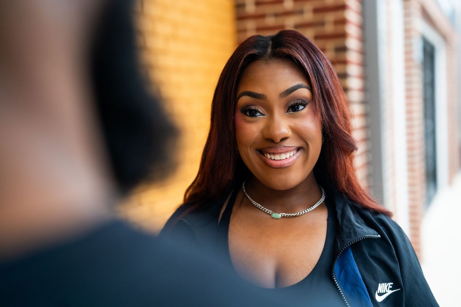 Smiling Black woman with red hair wearing Nike jacket against brick wall background.