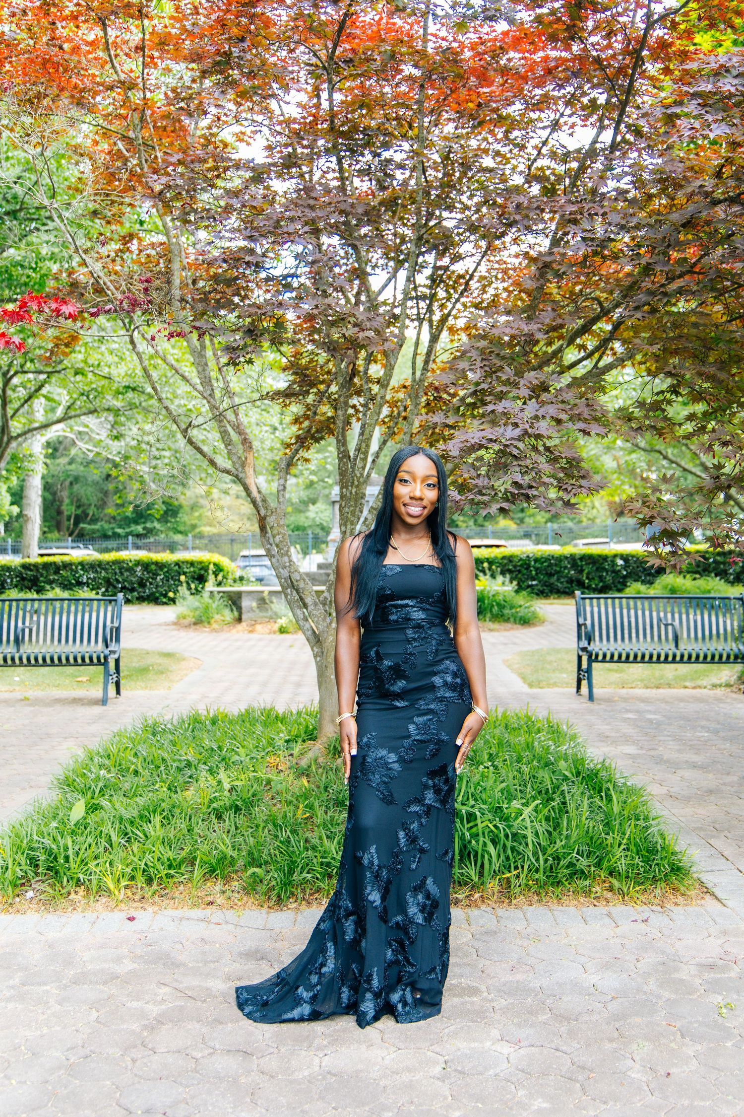 Woman in stunning black floral gown posing under colorful Japanese maple tree in park.