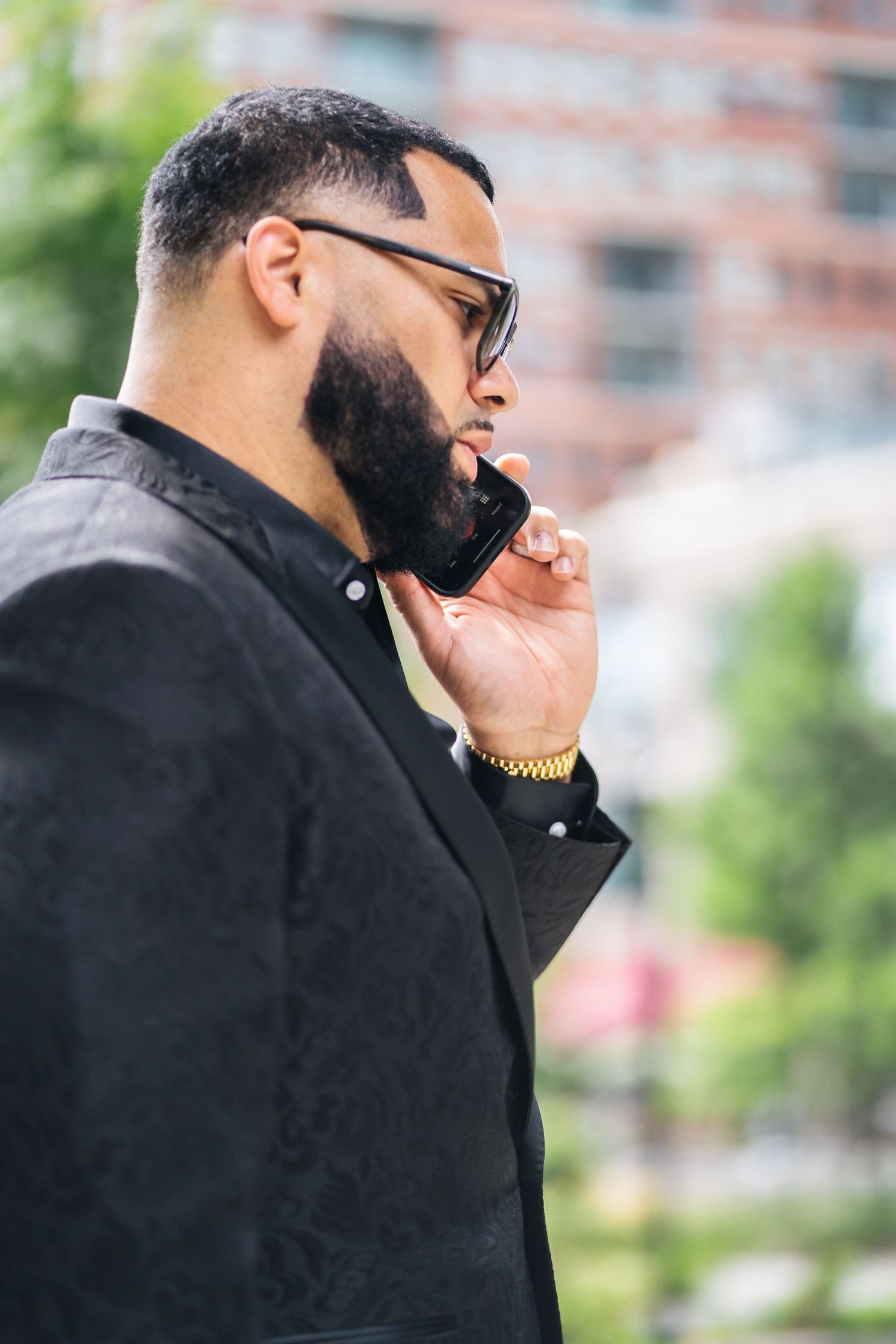 Stylish bearded Black man in black suit talking on smartphone outdoors with urban background.