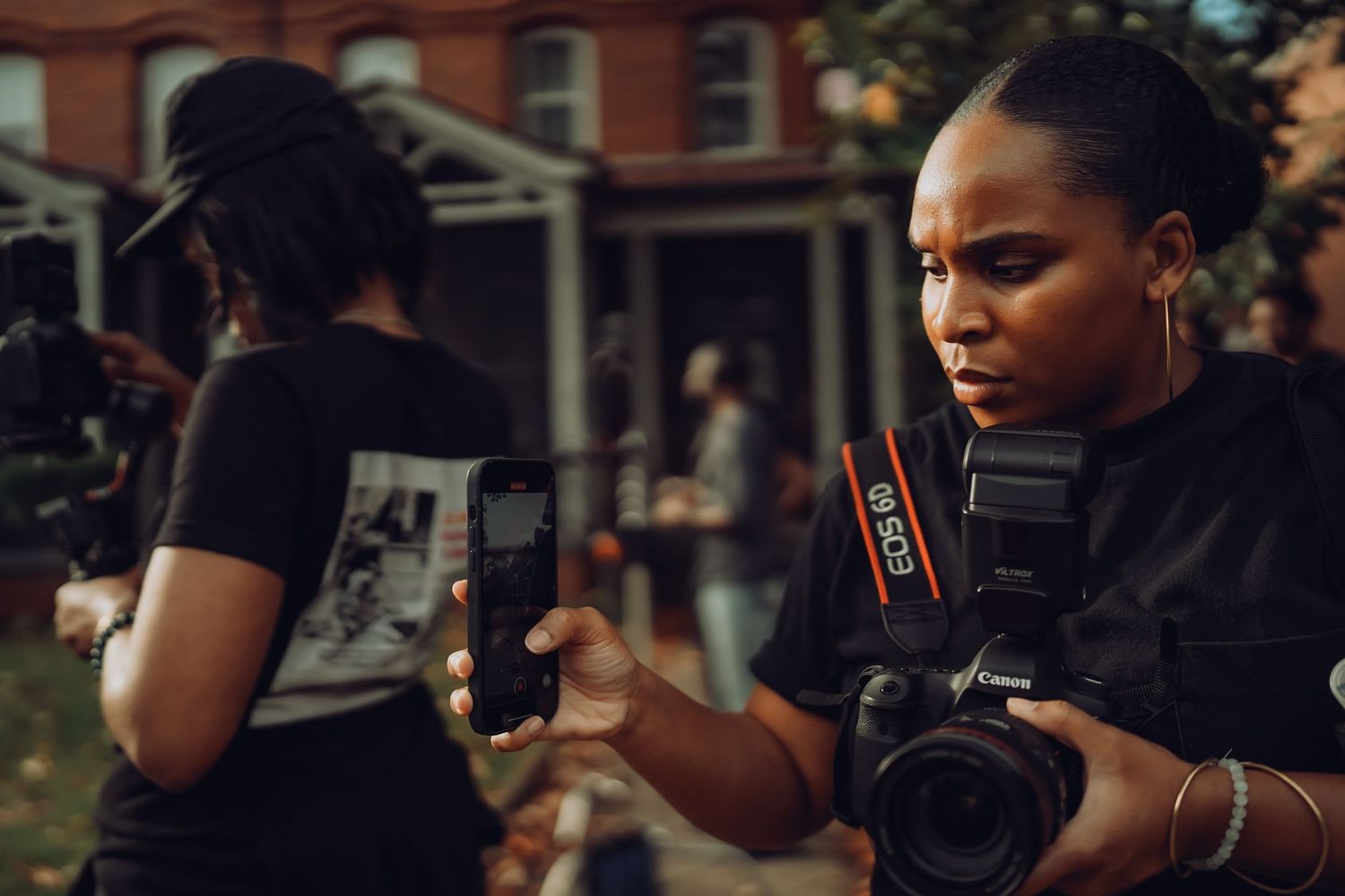 A female videographer holds a DSLR camera and smartphone on an outdoor film set.