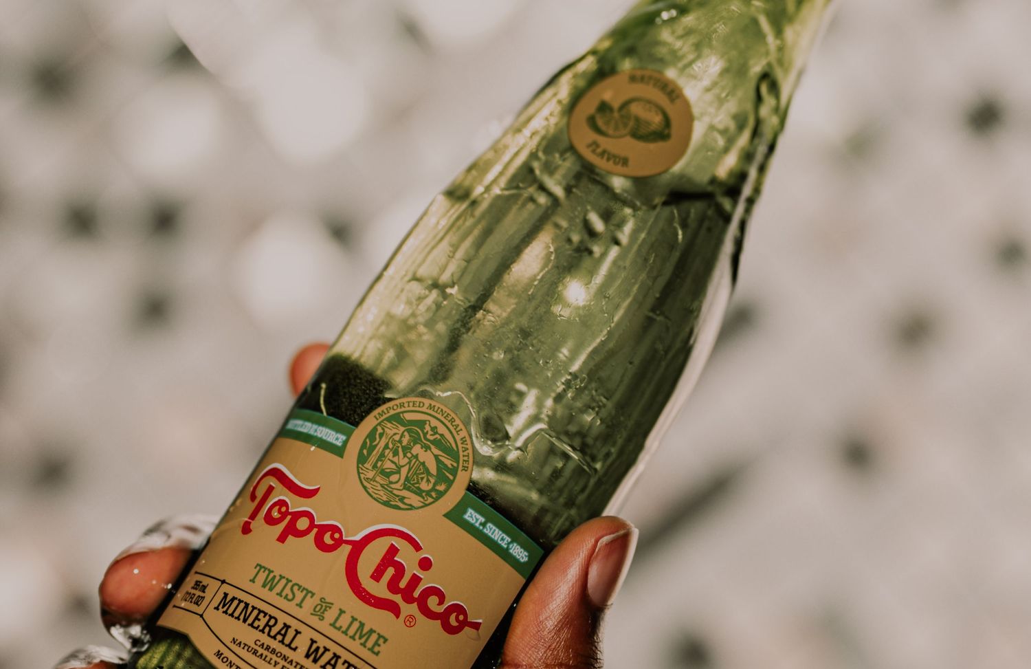 Hand holding a Topo Chico Twist of Lime mineral water bottle against a bokeh background.