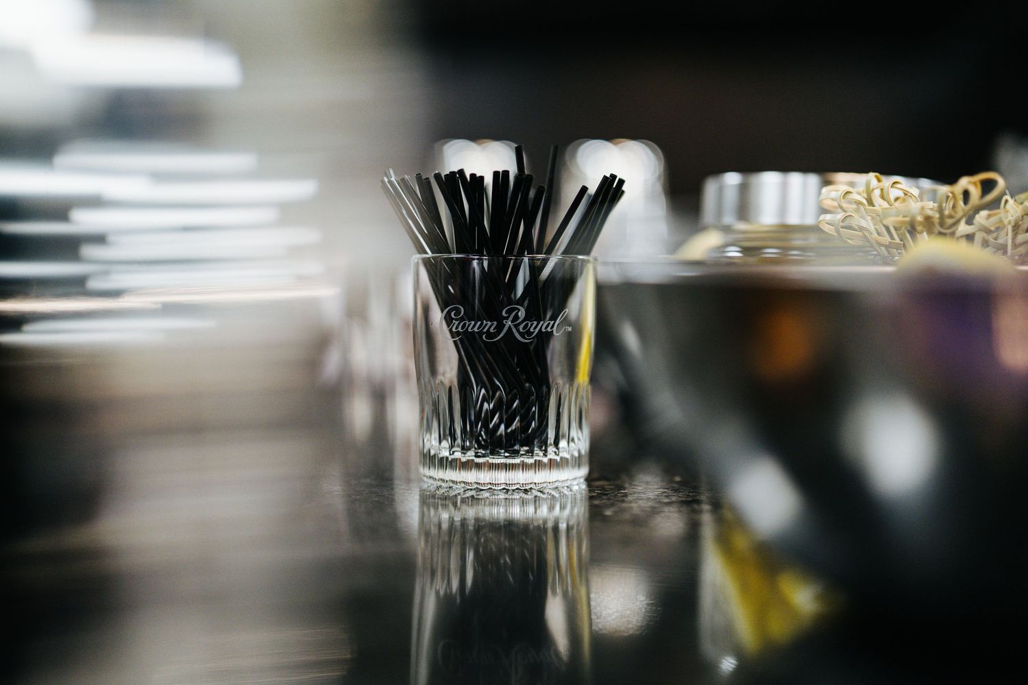 A Crown Royal branded glass filled with black cocktail straws sits on a reflective bar surface.
