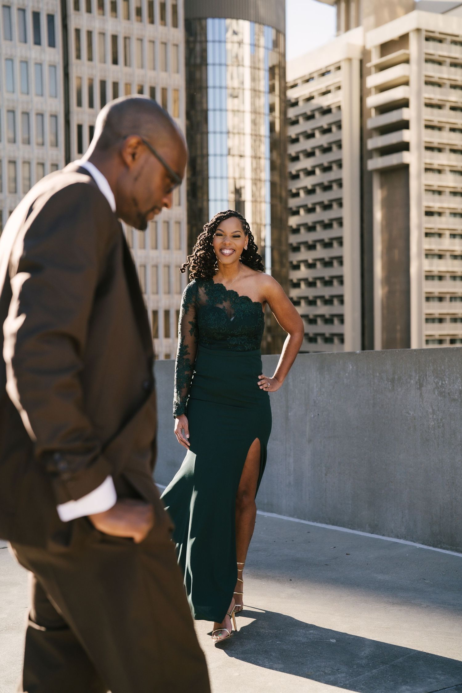 Elegant couple in formal attire posing on urban rooftop, woman in green gown smiling.