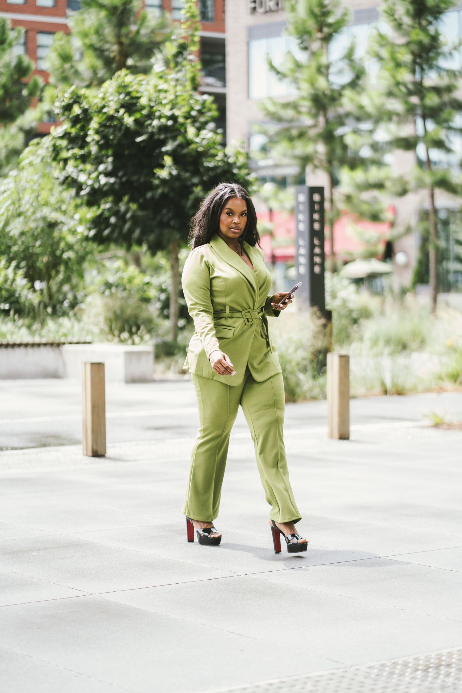 Stylish woman in green pantsuit walking outdoors on city sidewalk holding smartphone wearing platform heels.