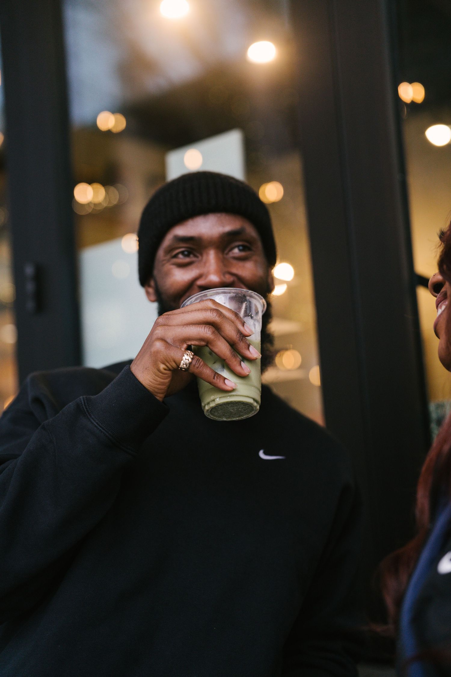 Black man in Nike beanie sipping iced drink outdoors with warm bokeh background.