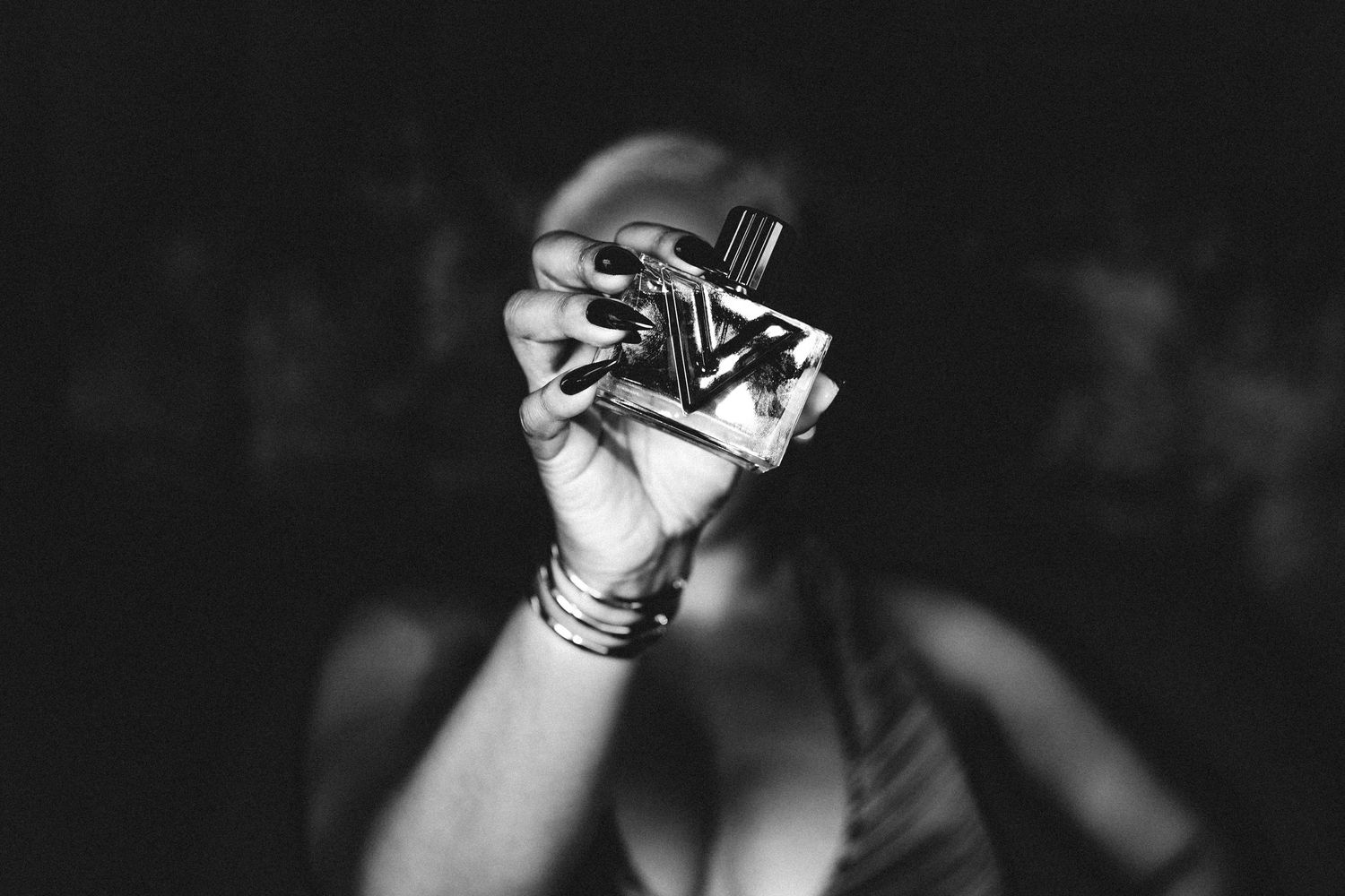 Black and white photo of a woman with stacked bracelets holding a perfume bottle up toward the camera in dramatic low light.