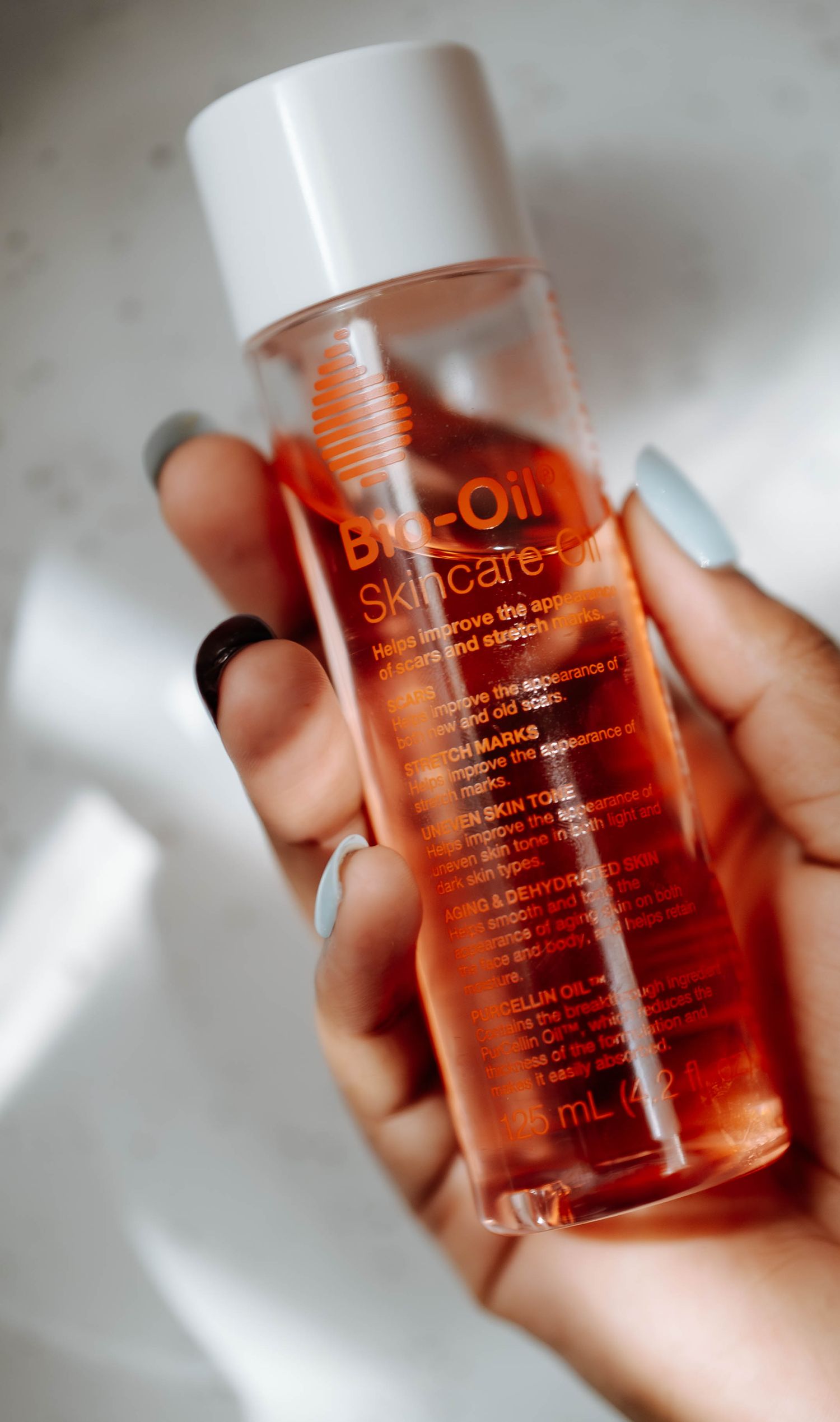 Hand with blue nails holding a Bio-Oil Skincare Oil bottle against a white background.