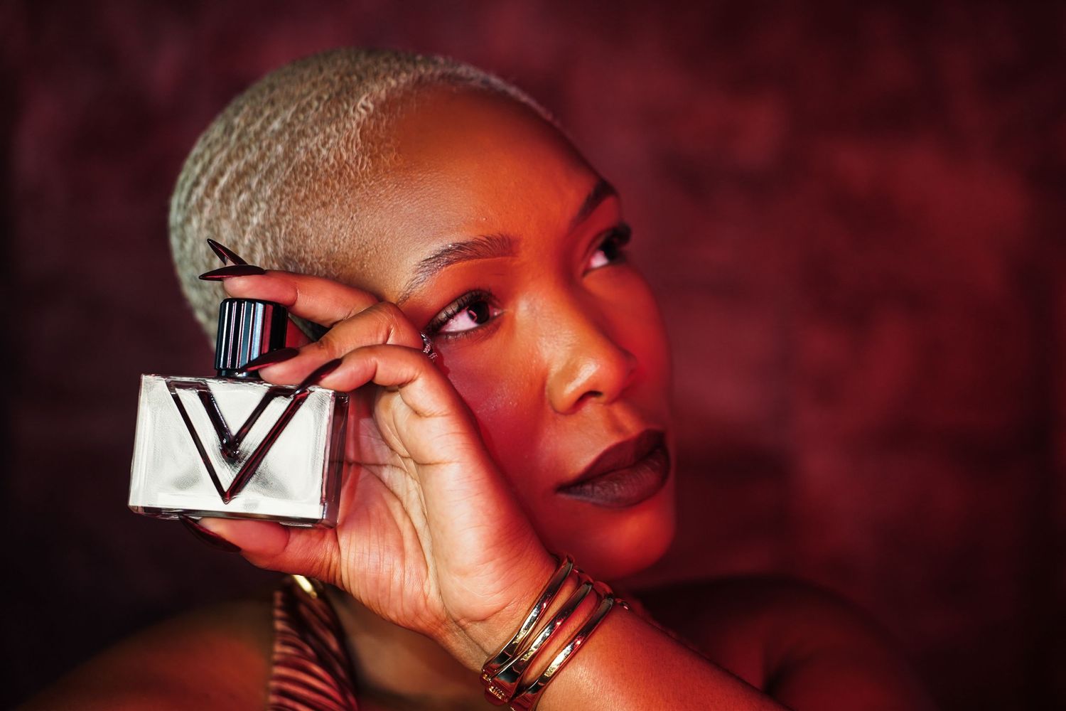 Woman with short platinum hair and bold lipstick holds a square perfume bottle with a V logo up to her eye against a red background.