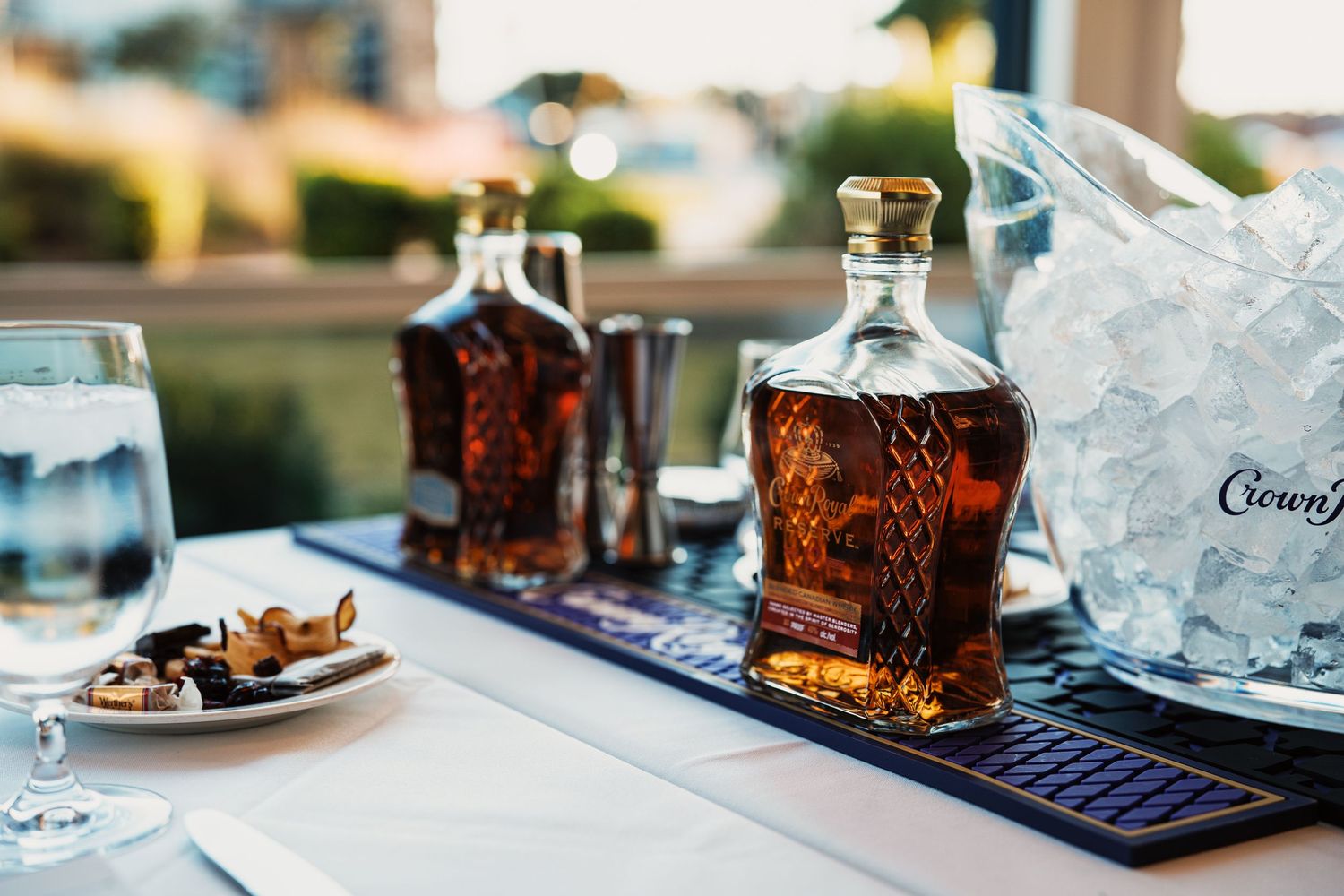 Crown Royal whiskey decanters displayed on a bar mat beside an ice bucket at an elegant outdoor event.