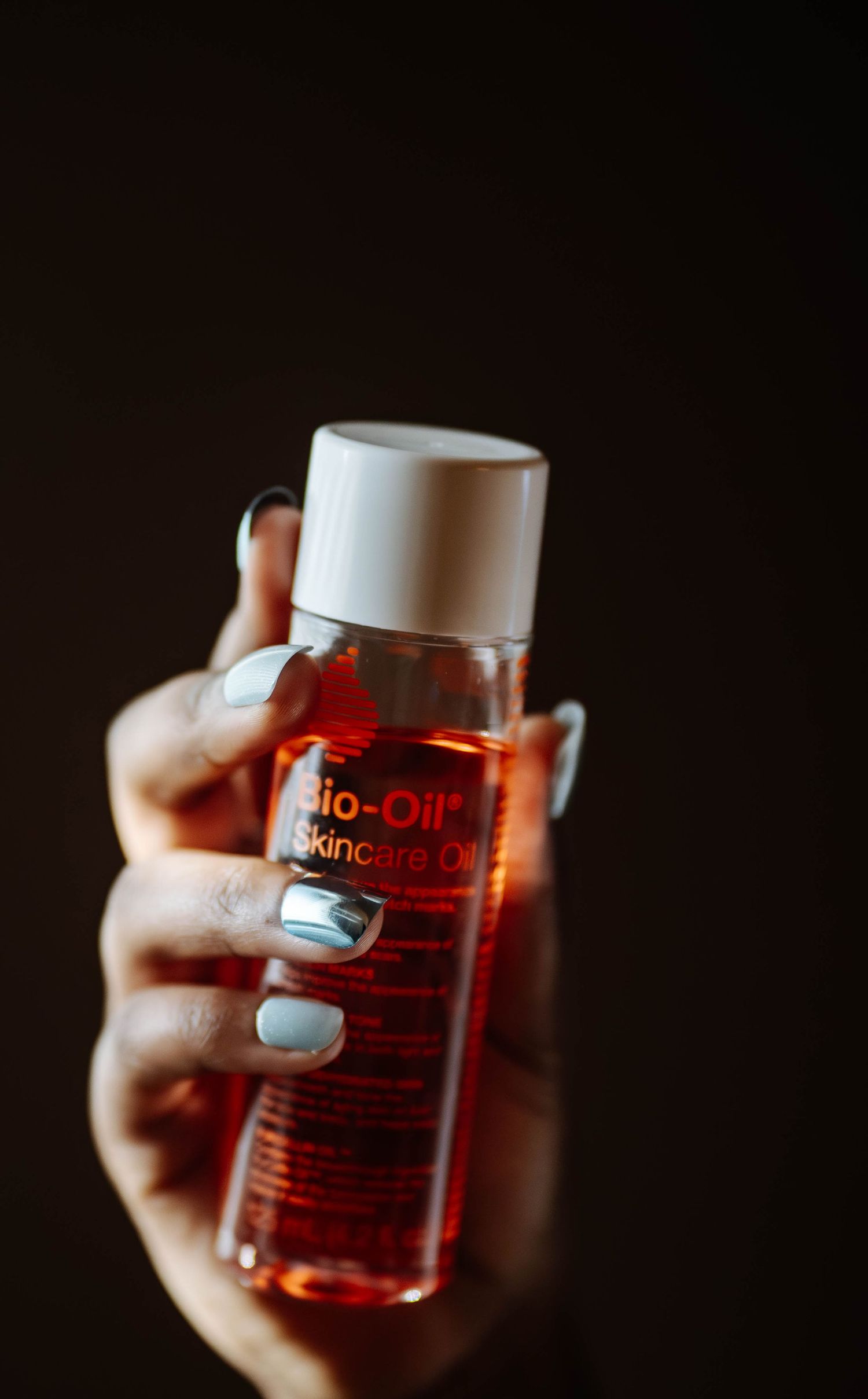 Hand with light blue nails holding a Bio-Oil Skincare Oil bottle against a dark background.
