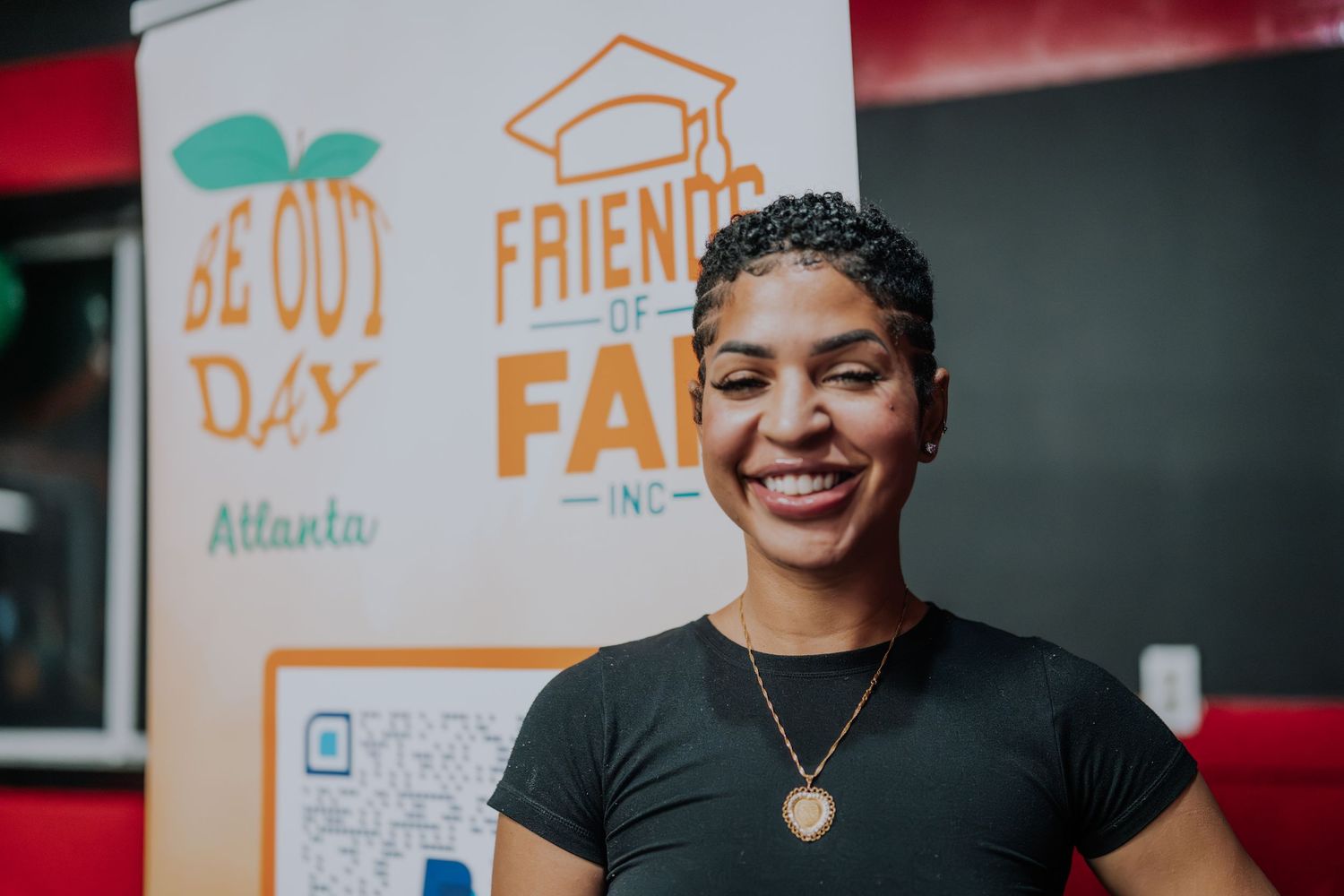 Smiling woman in black shirt stands before Friends of FAM Inc. and Be Out Day Atlanta banners.
