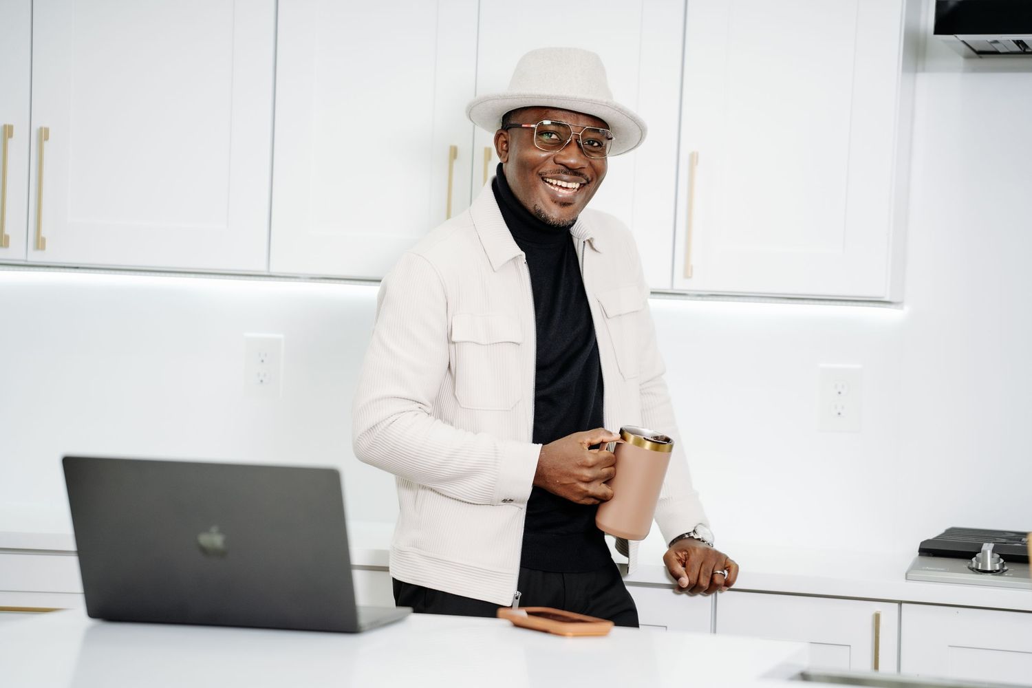 Stylish man in white hat holding cup standing by laptop in kitchen.