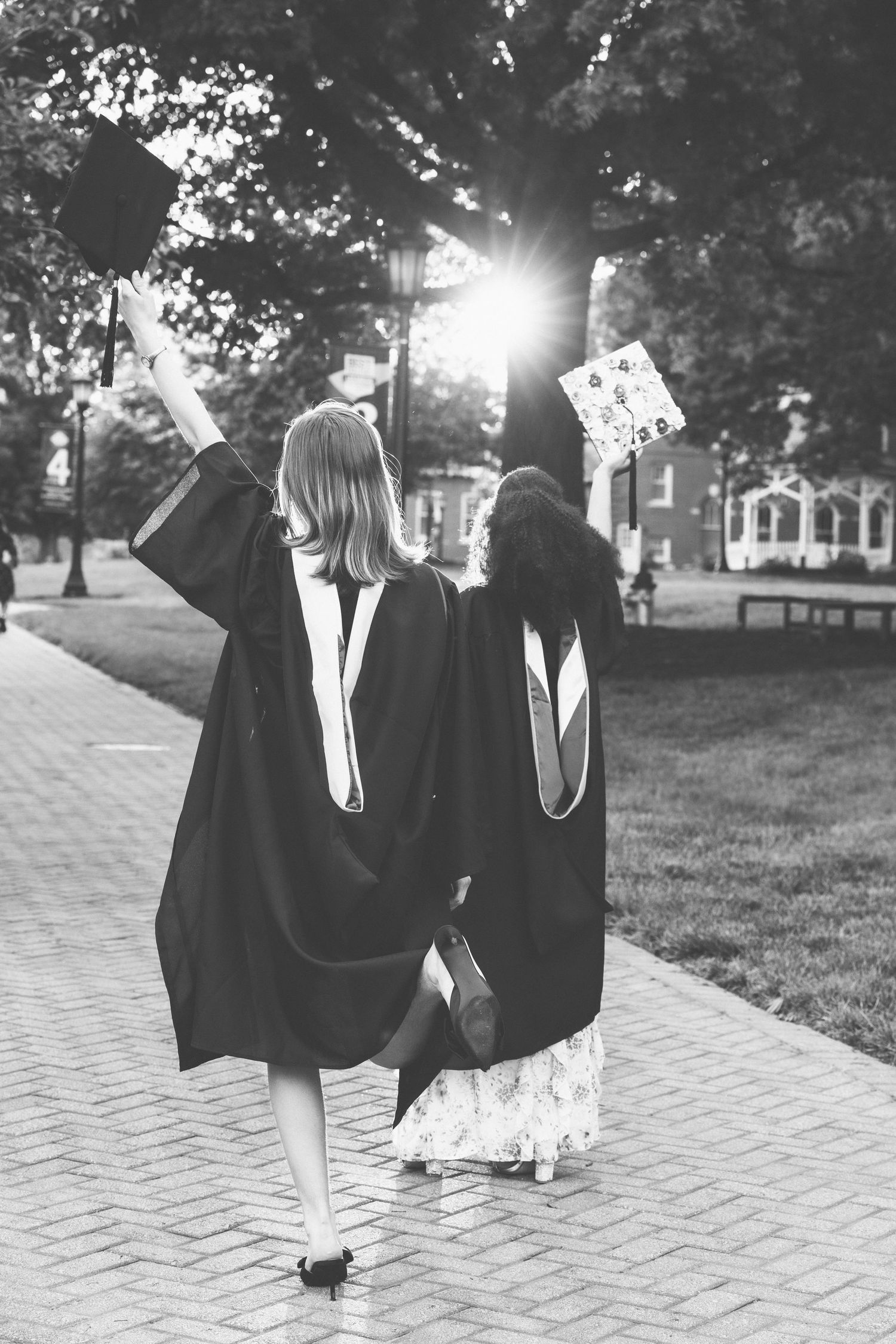Two graduates in black robes walk away tossing caps on brick campus path.