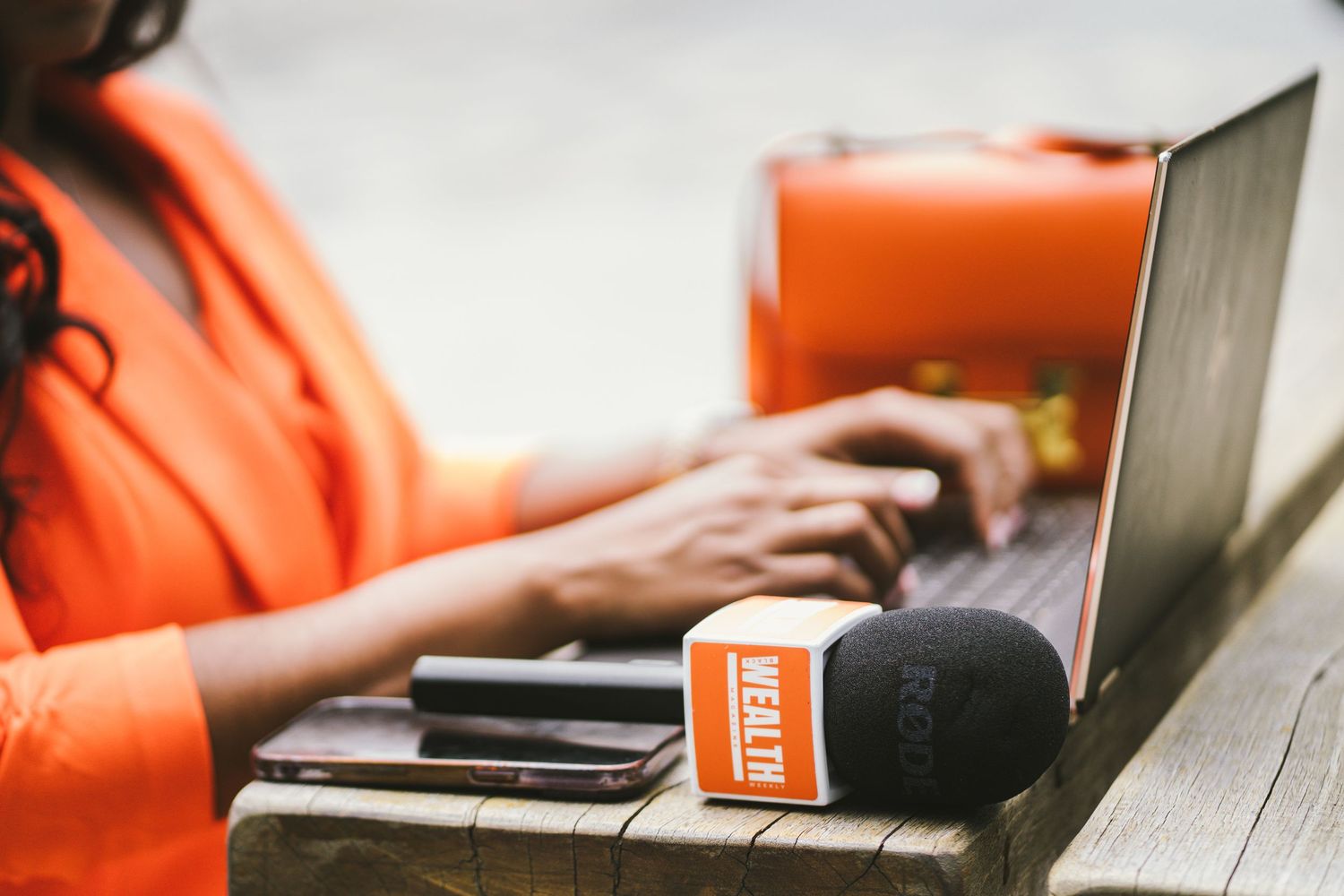 Close-up of hands typing on laptop with branded microphone and orange bag on wooden table outdoors.