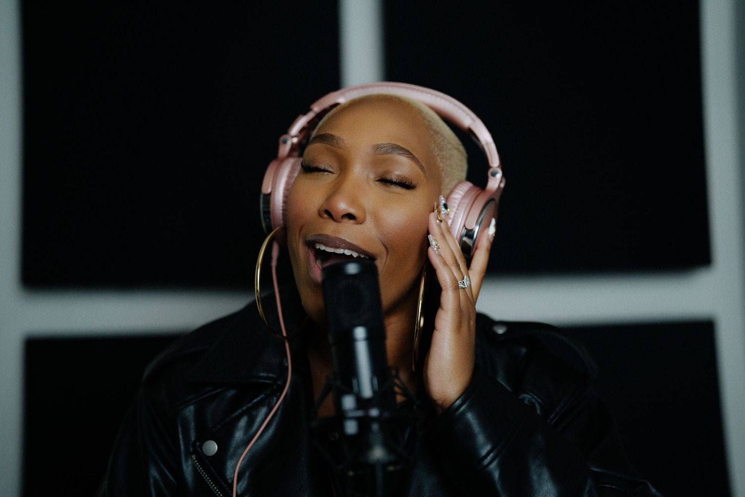 Woman singer recording in studio wearing pink headphones and black leather jacket with eyes closed at microphone.