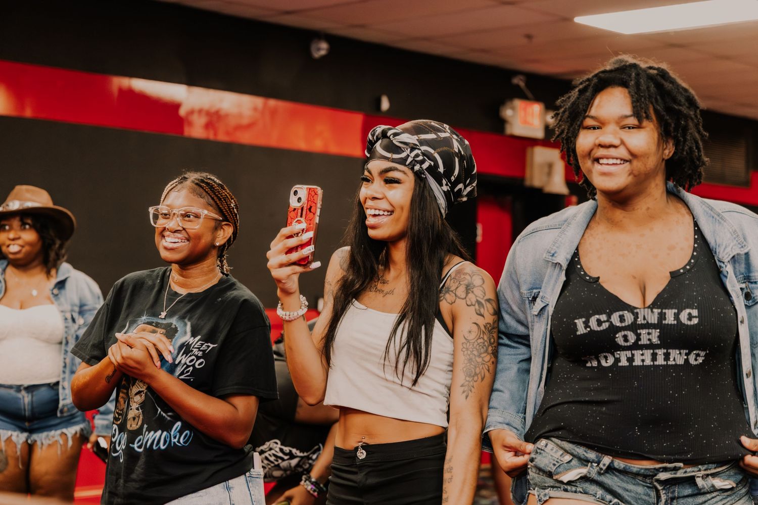 Three women laugh and celebrate together at a lively indoor event, one wearing an Iconic t-shirt.