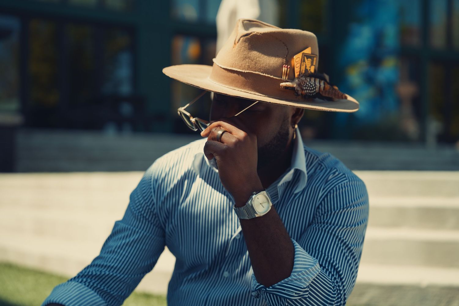 Fashionable Black man in striped shirt and brown fedora hat adjusting sunglasses outdoors.