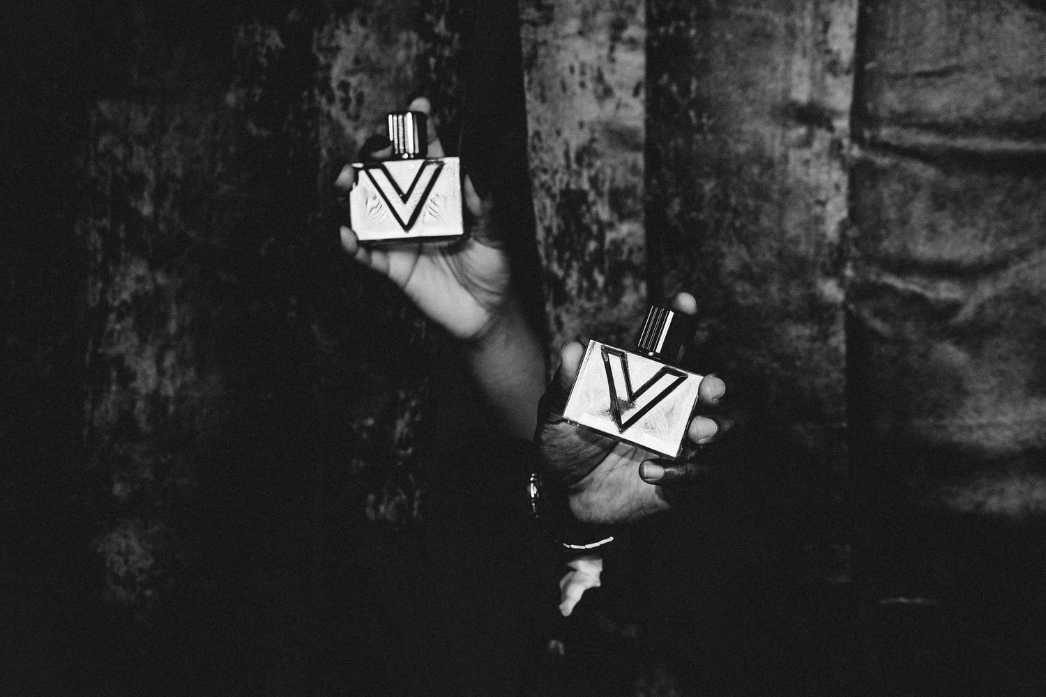 Black and white photo of two hands each holding a square perfume bottle with a V logo in a dark, moody setting.