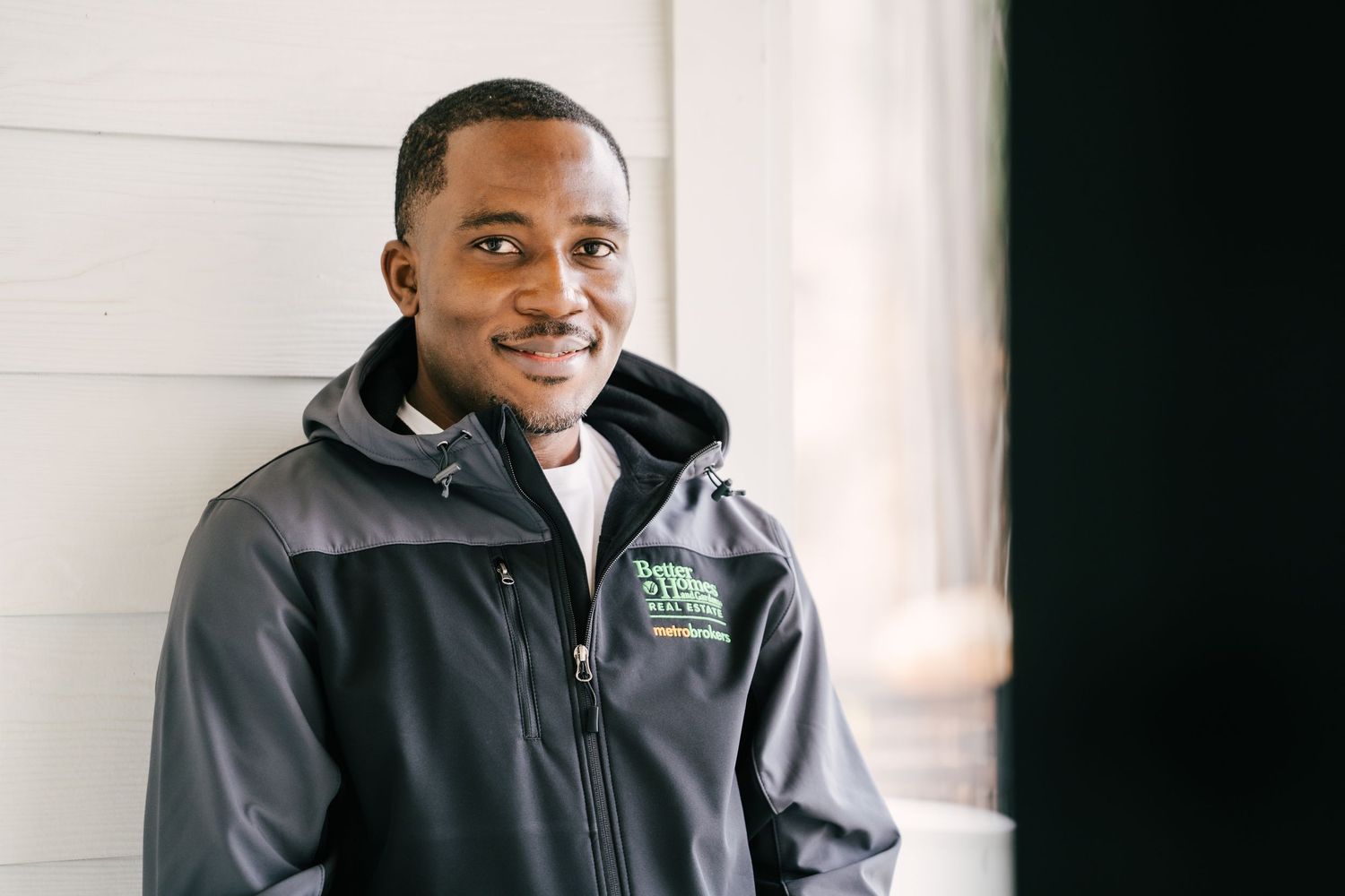 Young man in gray zip-up jacket smiling against white wall.