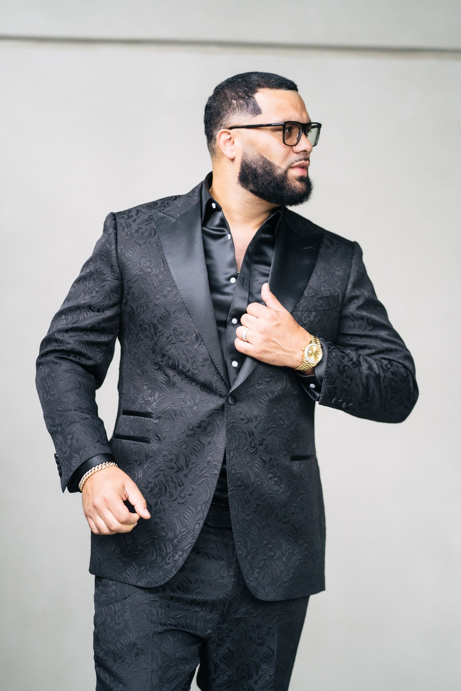 Well-dressed bearded man in black floral tuxedo suit with gold watch posing against gray wall.