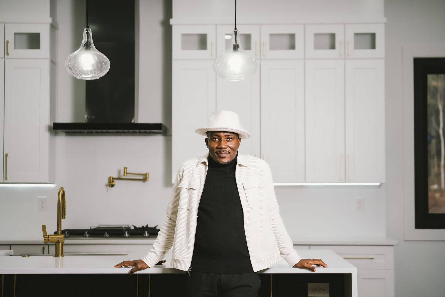 Stylish Black man in white hat and jacket leaning on kitchen island counter.