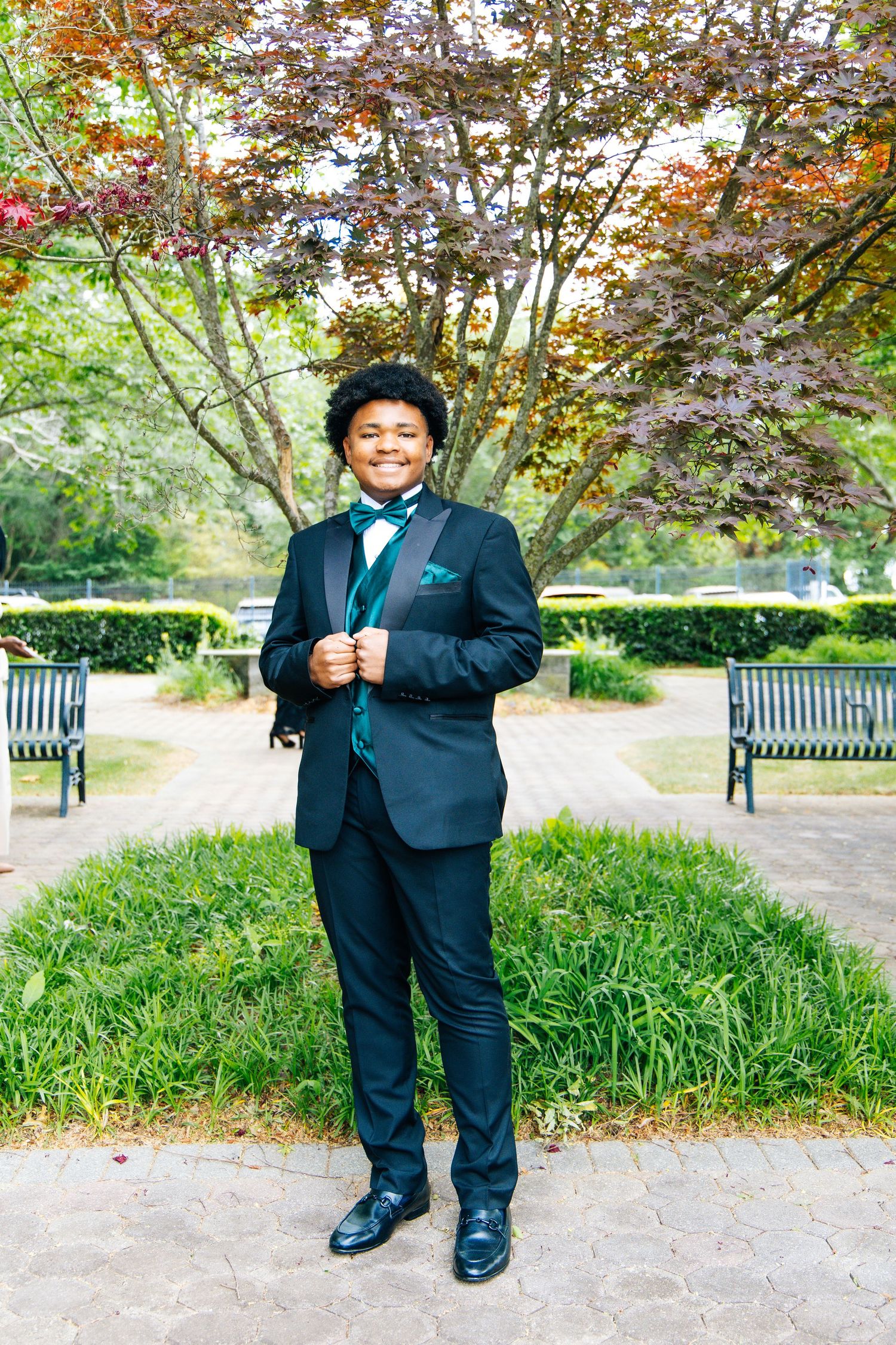 Young man in black suit with teal bow tie and vest smiling in park.
