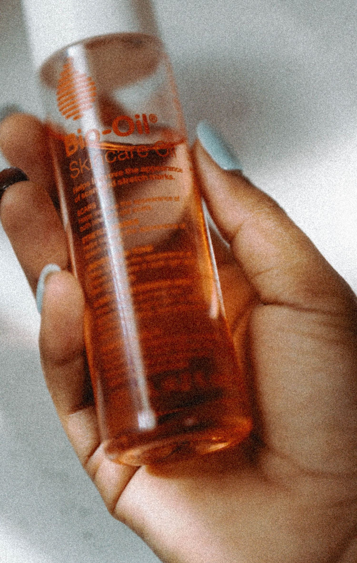 Hand holding a translucent orange Bio-Oil Skincare Oil bottle with light blue manicured nails.