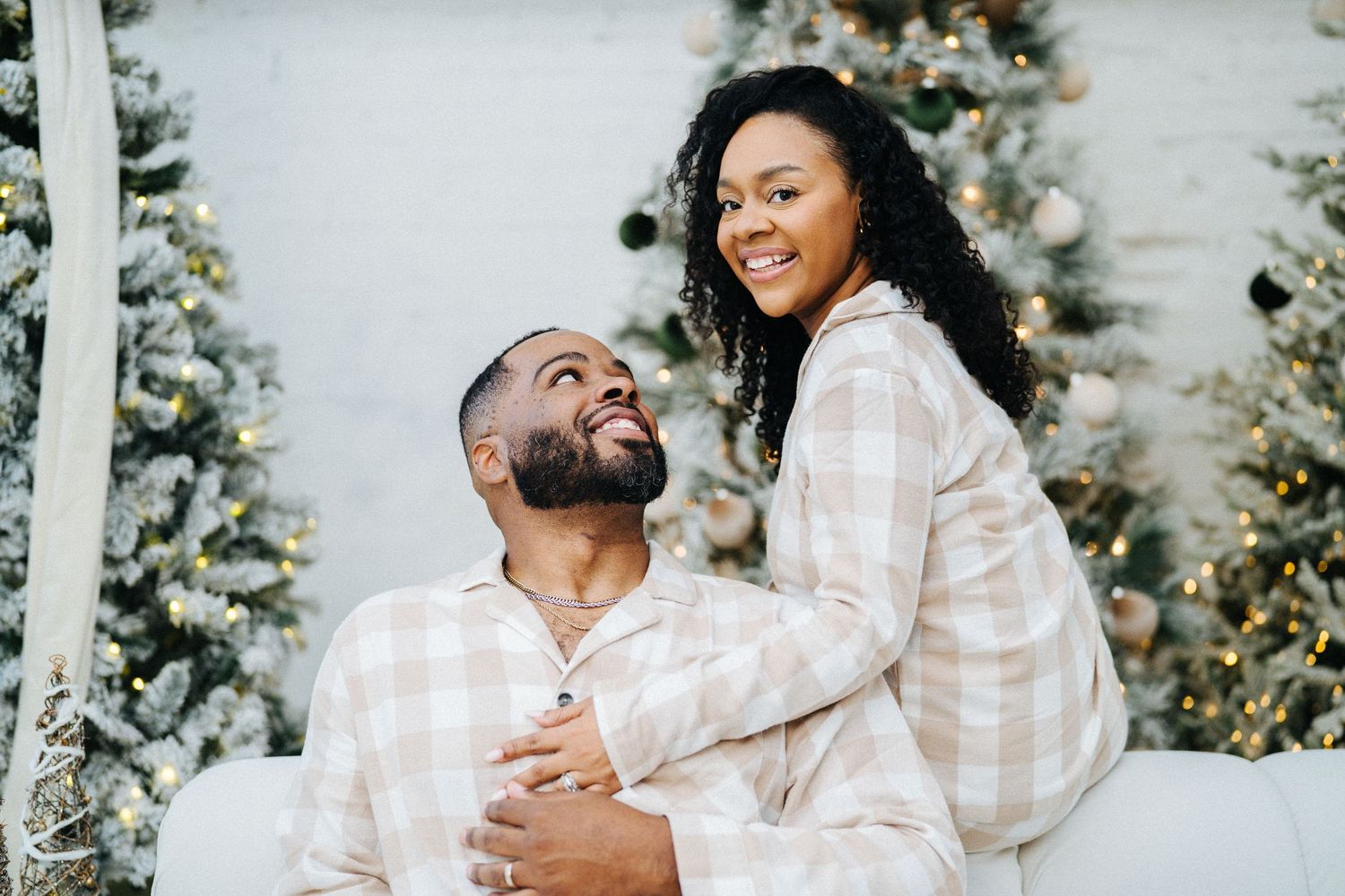 Happy Black couple in matching plaid pajamas embracing near decorated Christmas trees.