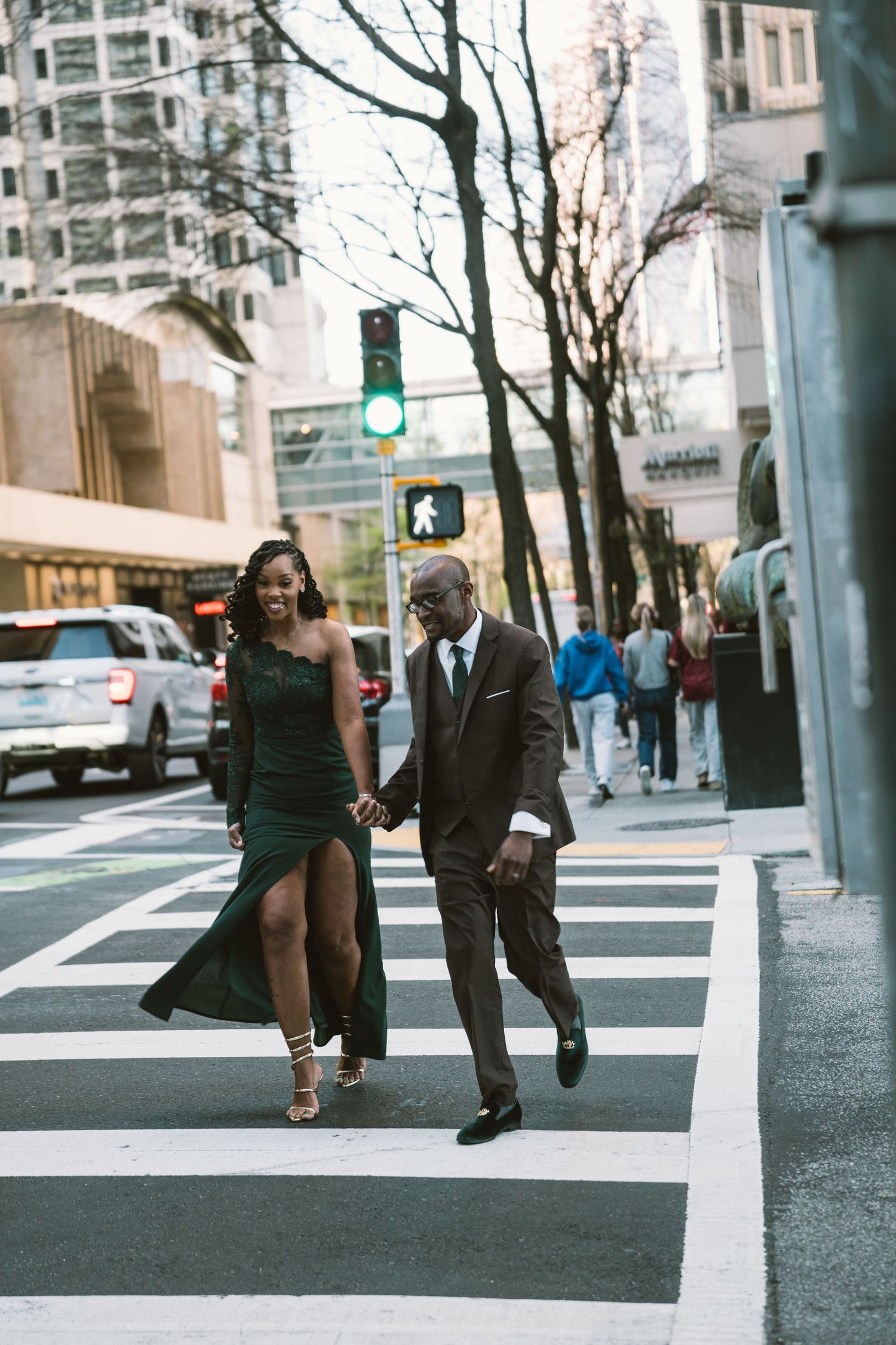 Elegant couple holding hands while crossing a city street, woman in green gown and man in dark suit.