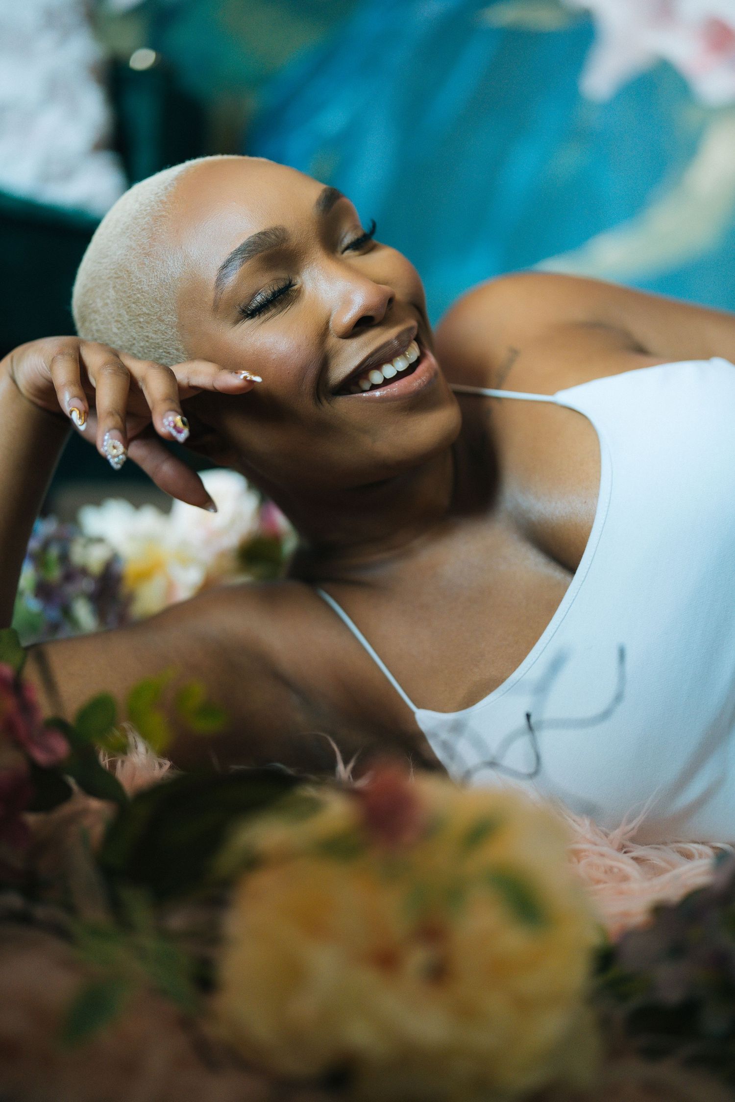 A joyful woman with a shaved head in a white dress lying among colorful flowers.