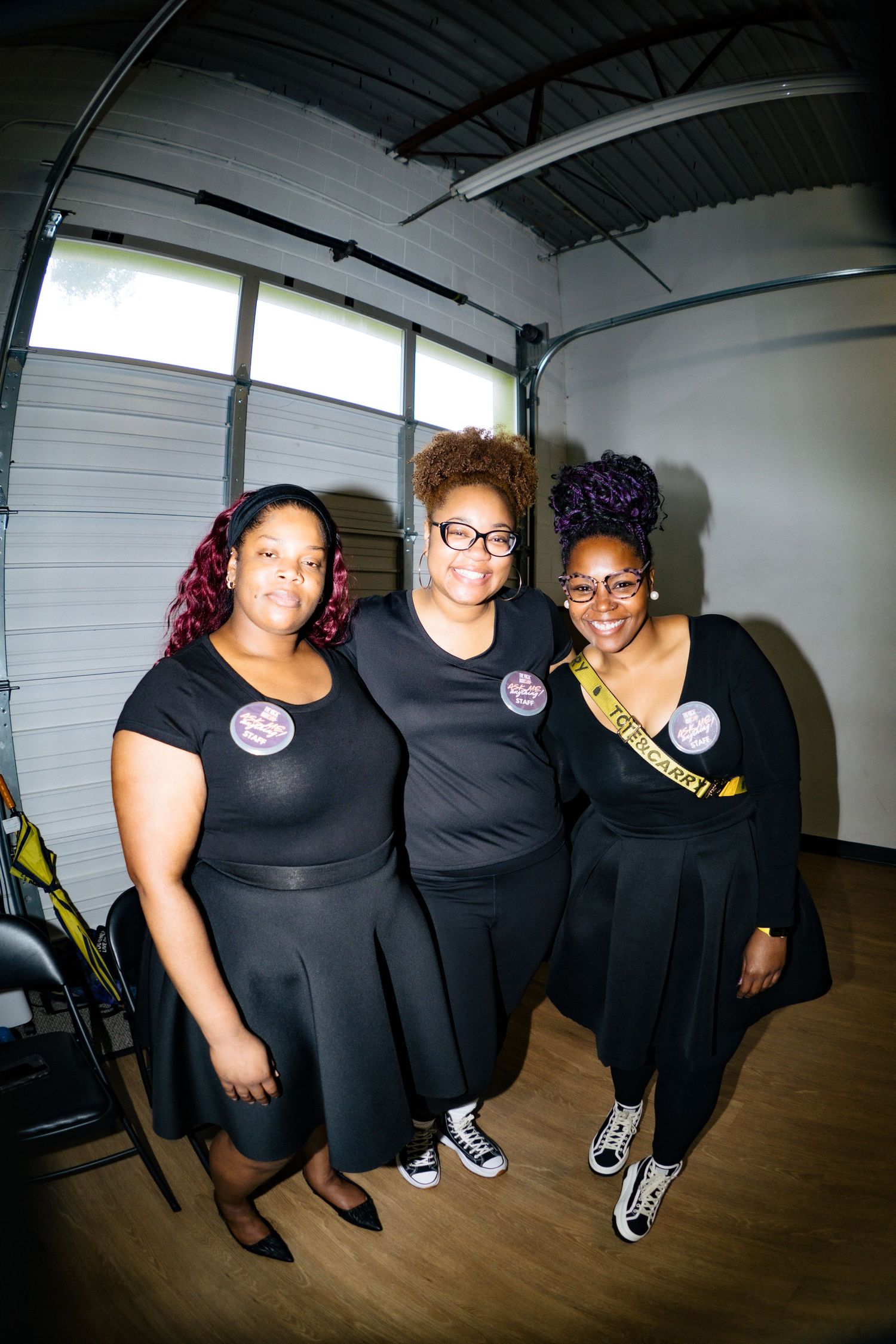 Three women in matching black outfits smile together at an indoor event, one wearing a gold sash.