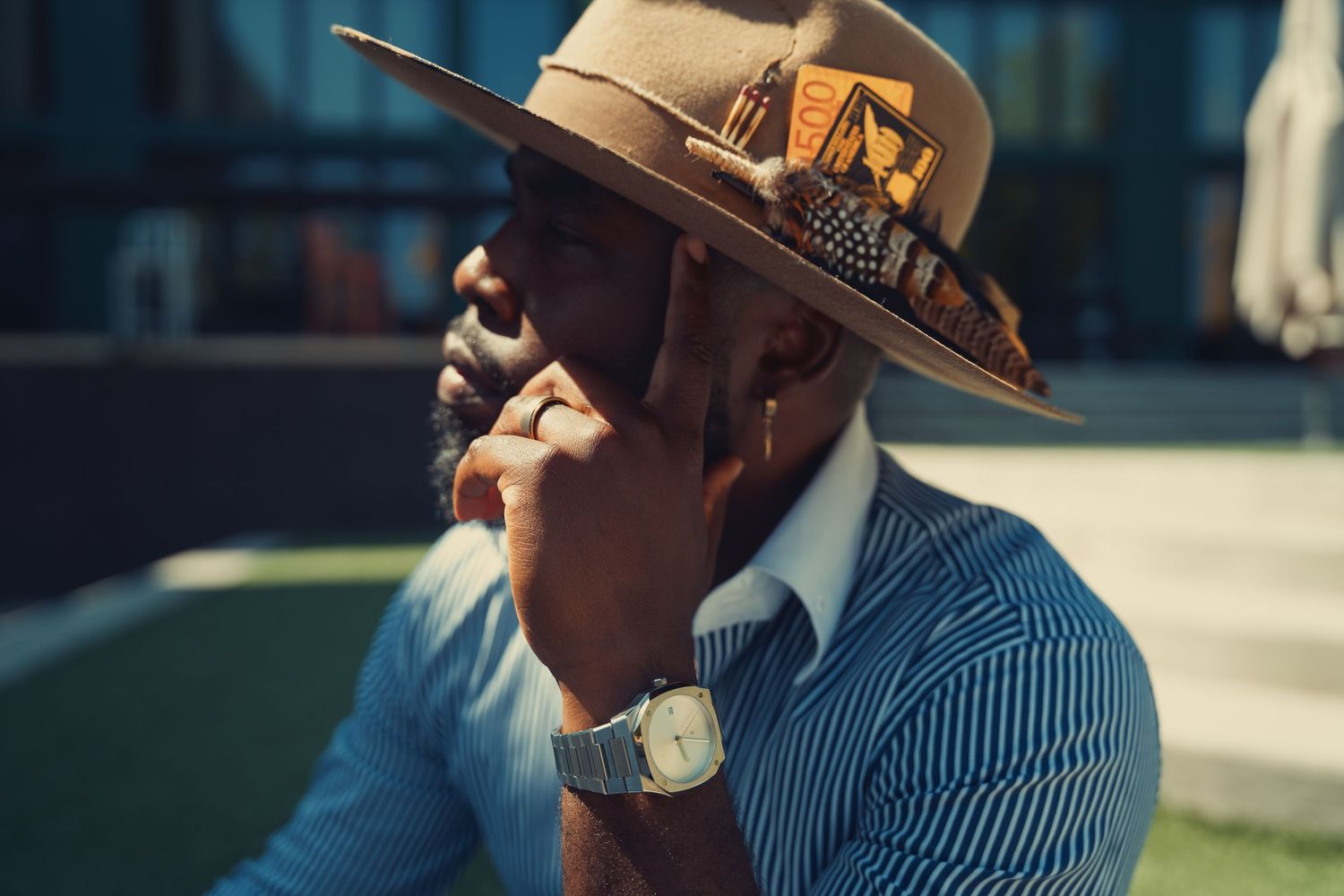 Thoughtful Black man wearing brown wide-brim fedora hat and striped shirt with gold watch.
