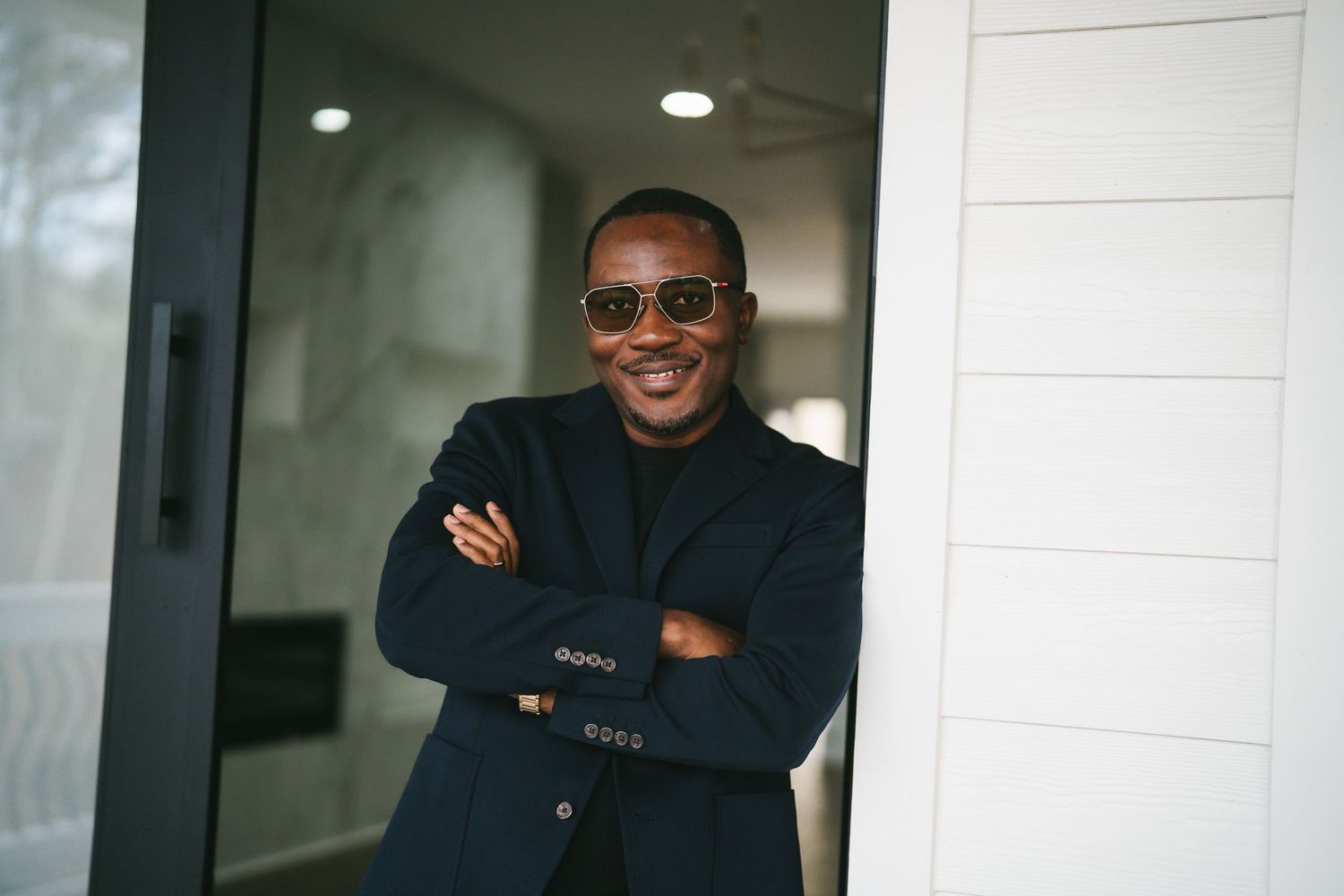 Man in black jacket with sunglasses smiling with arms crossed outdoors.
