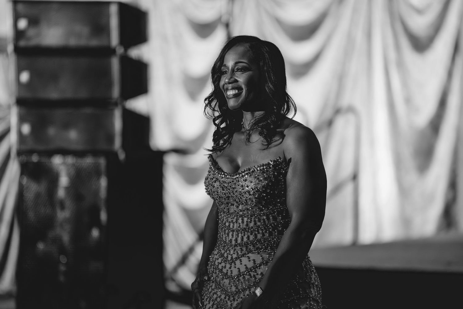 Woman in elegant beaded gown smiling warmly on stage in black and white concert photography.
