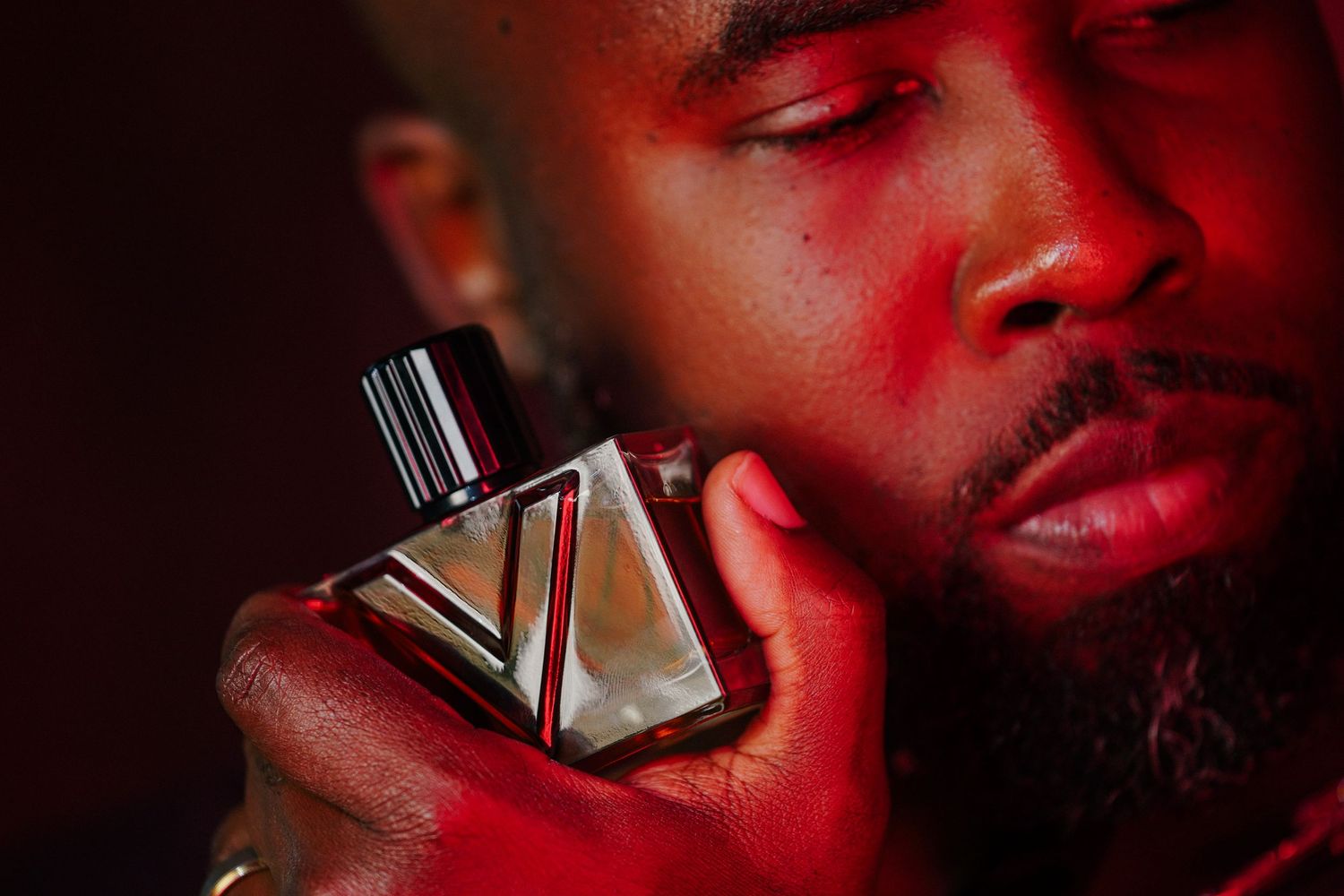 Bearded man with eyes closed holds a sleek angular cologne bottle against his cheek, bathed in moody red lighting.
