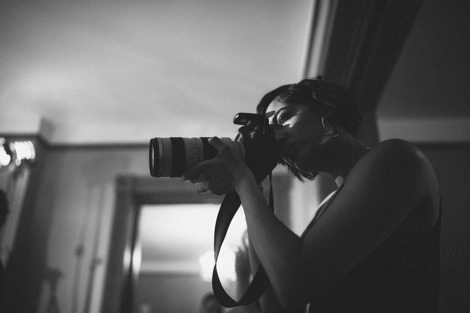 Black and white photo of a photographer aiming a DSLR camera with a telephoto lens indoors.