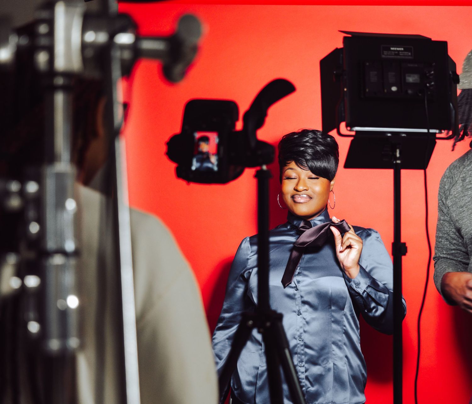 A woman in a satin shirt poses confidently in front of cameras on a red background.