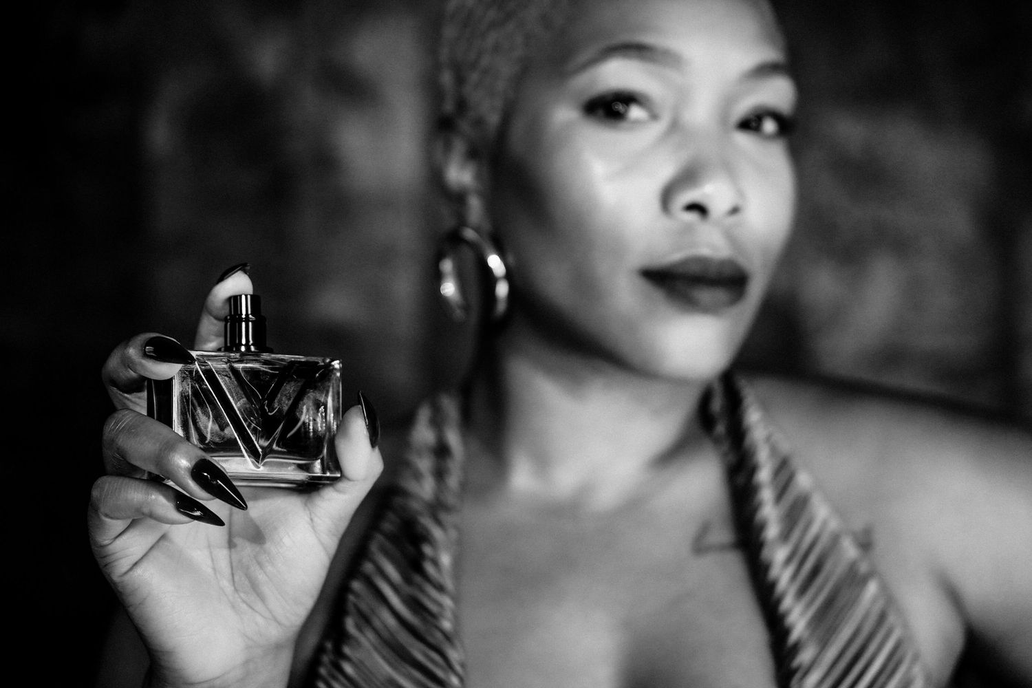 Black and white portrait of a woman with short hair and hoop earrings holding a glass perfume bottle toward the camera.